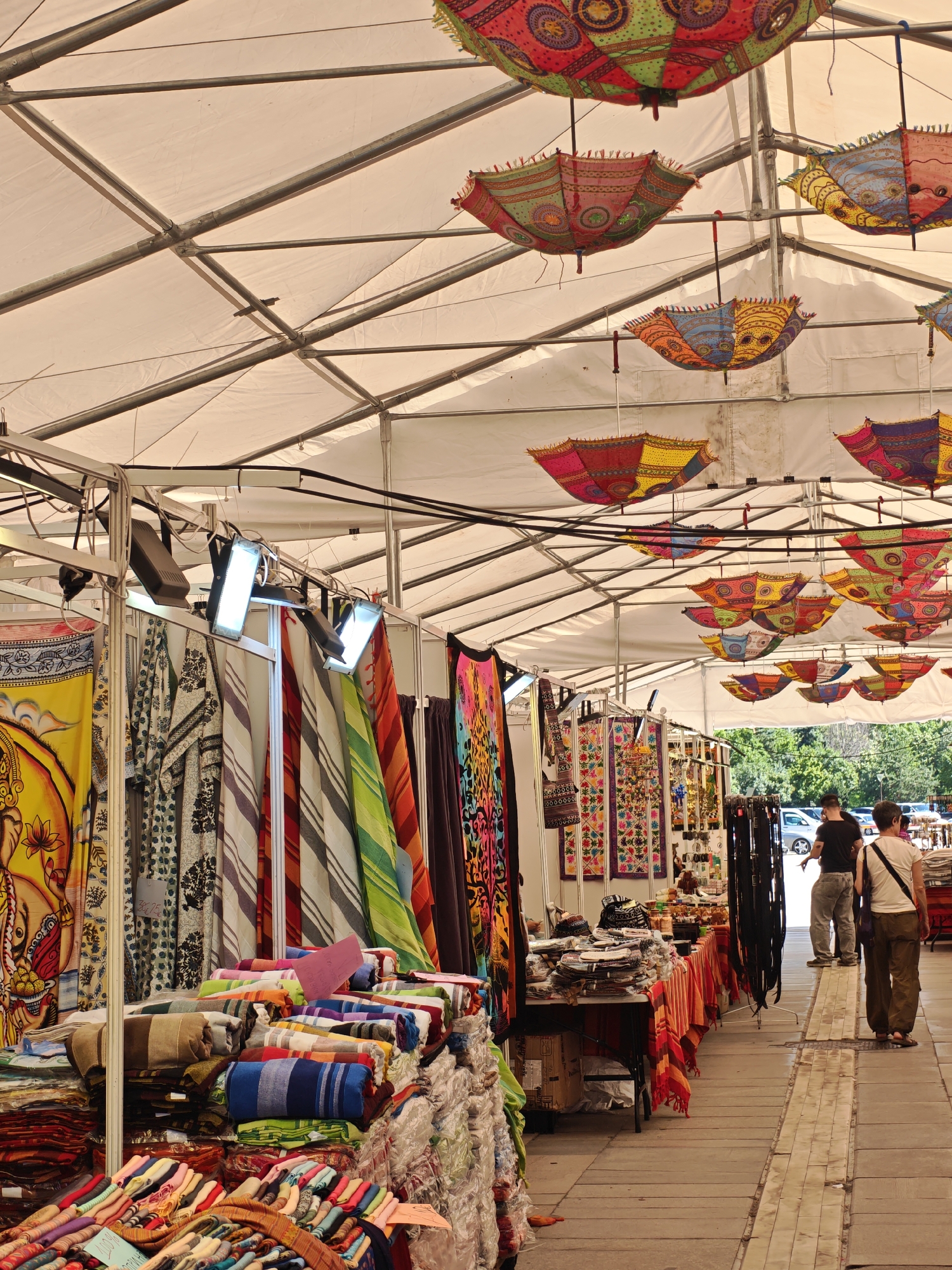 This image depicts an outdoor market stall beneath a large, white tented canopy, likely in Sofia, Bulgaria, given the context provided. The canopy is supported by a metal frame. Numerous colorful, patterned umbrellas are suspended from the canopy's structure, adding to the vibrant atmosphere.
The stall itself is filled with textiles, including what appear to be scarves, blankets, and fabric hangings, displayed on tables and racks. Several people are visible in the background, browsing the stalls. Two individuals, a man and a woman, are walking away from the camera. The man is wearing a black t-shirt and gray pants, while the woman is in a white t-shirt and khaki pants. They appear to be customers. The ground is paved with tiles, and trees and a parking lot with cars are visible beyond the market area. The lighting suggests it is daytime.
