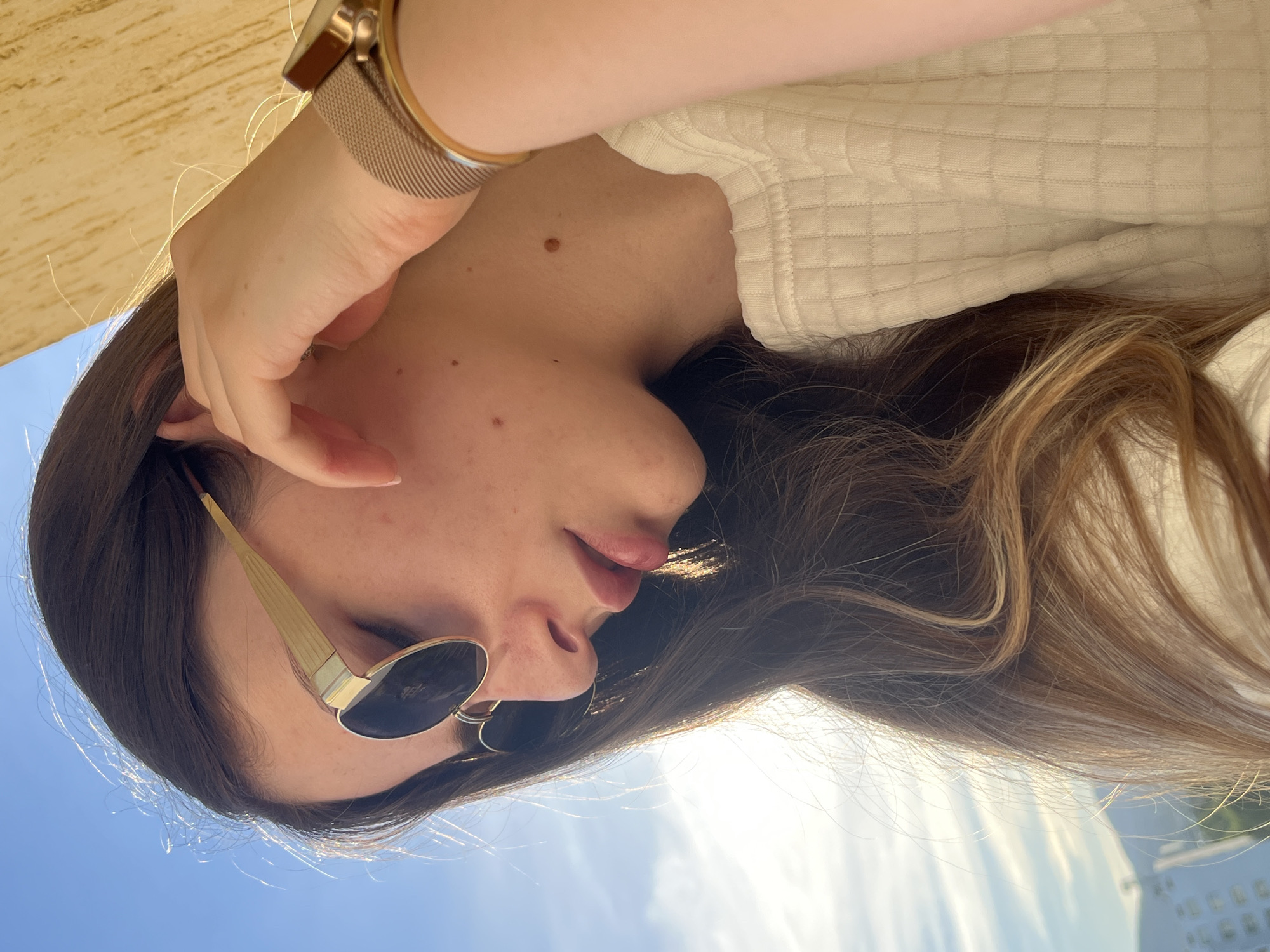 A close-up, over-the-shoulder shot captures a young woman from the neck up, as she looks down and to her left. She has long, dark brown hair with lighter highlights that fall around her face and shoulders. She is wearing round, gold-rimmed sunglasses with dark lenses, and her right hand is raised to her ear, fingers gently touching her temple. A gold mesh watch with a rose gold casing is visible on her left wrist. She is wearing a cream-colored, textured shirt. The background is a bright blue sky with thin, wispy clouds, and a hint of a light-colored building or structure is visible in the upper right corner, casting a shadow that creates an awning-like effect. The overall lighting suggests it is daytime, possibly late afternoon, with the sun providing a warm glow.
