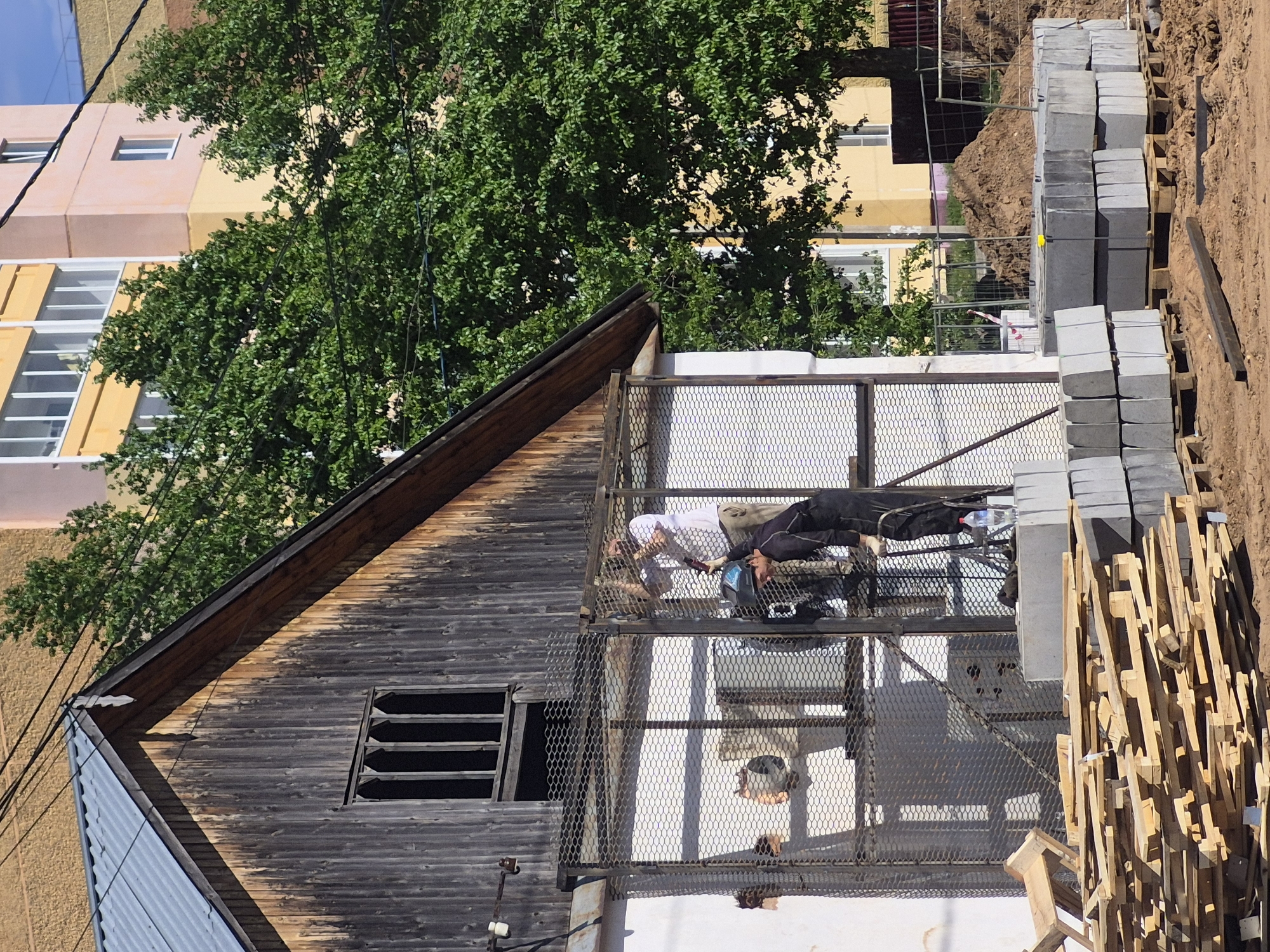The scene presents a combination of old and new architecture in what appears to be an urban environment. An old, dark wooden building with a weathered roof and a barred window is positioned to the left. Adjacent to it is a modern addition with a metal mesh balcony structure. On the balcony, two individuals appear to be resting, one in light-colored clothing and the other in dark clothing.
To the right of the building, there is construction material including stacks of gray blocks and wooden pallets. In the background, a multi-story building with a mix of tan and peach-colored facades is visible, partly obscured by a green tree. Above, a clear blue sky is present. The ground in the foreground has a sandy, dirt-like appearance.
