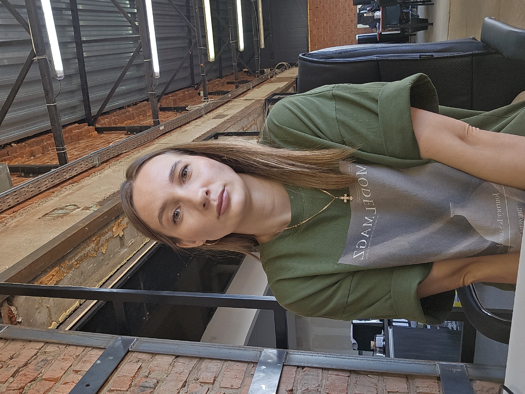 A young woman is in an industrial-style setting. She is wearing an olive green shirt with a graphic print and a gold necklace with a cross pendant. The background shows a mix of exposed brick walls, metal beams, and fluorescent lights typical of an industrial building. The location is Chaikovsky, Russia. She appears to be sitting in a chair, possibly in a salon or studio.