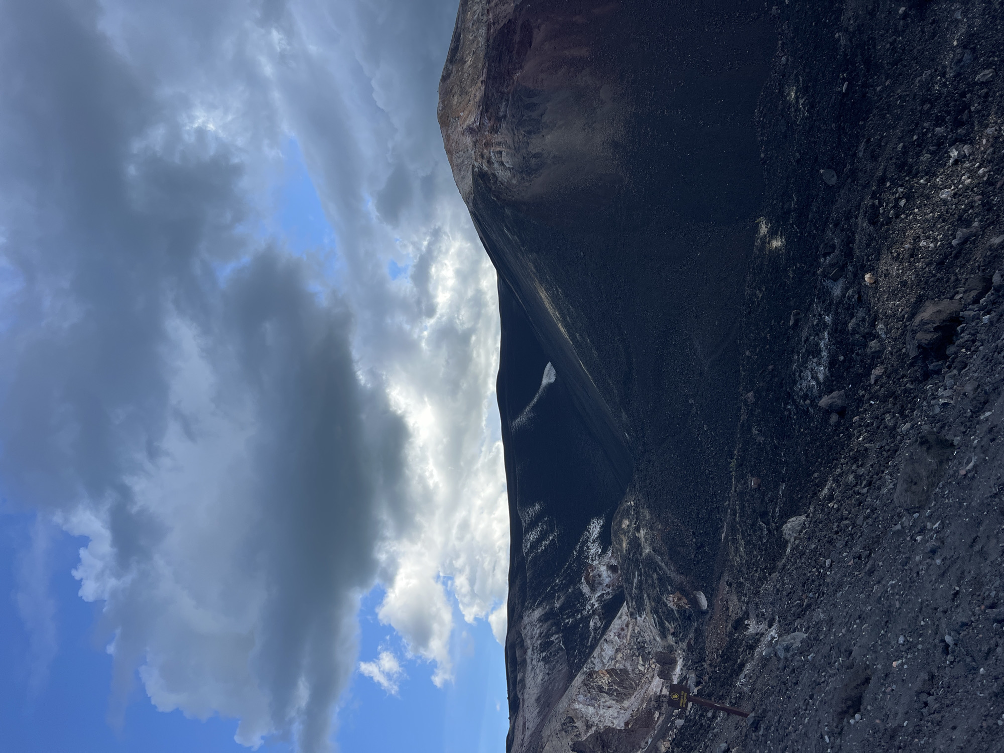 The image depicts a scene with a large, dark, rocky formation that takes up the majority of the right side. The formation appears to be a cliff face or a massive pile of dark, granular material mixed with rocks. The surface has a textured appearance, with varying shades of black and gray. There are some lighter-colored mineral deposits or lighter rock streaks visible on the lower portion of the formation. A small yellow sign with some text is visible near the bottom right corner of the formation.
To the left of the dark formation, the sky is visible with a mix of clouds and patches of blue sky. The clouds are mostly white and gray, with some appearing dense and heavy, suggesting potentially overcast conditions or the approach of a storm. The lighting indicates it is daytime, possibly late afternoon or evening due to the contrast between the bright sky and the dark formation, but this is not definitively ascertainable.
The scene appears to be an outdoor environment, possibly a quarry, volcanic area, or a large industrial site given the nature of the material. There are no people visible in the image. The overall mood conveyed is somewhat dramatic due to the stark contrast between the dark, imposing rock and the dynamic sky. The orientation of the image is such that the sky is on the left and the rocky formation is on the right, with the ground or base of the formation at the bottom.