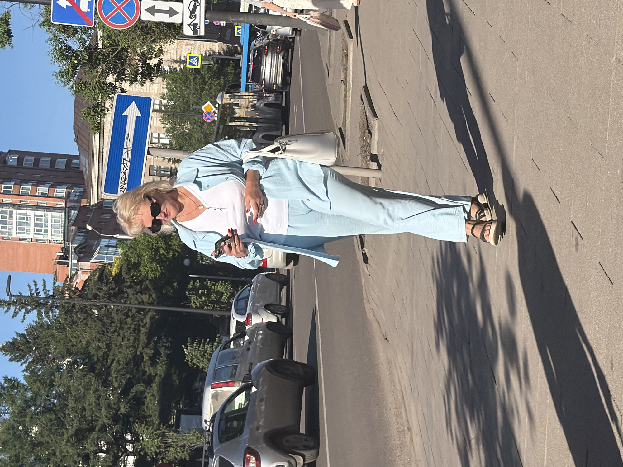 A woman in a light blue suit and white top is walking on a sidewalk in Krasnoyarsk, Russia. She is wearing sunglasses and holding a smartphone. She has a white purse slung over her shoulder. In the background, there are parked cars, trees, and buildings. Several traffic signs are visible, including one with a white arrow pointing upwards. The image appears to have been taken during the day, as indicated by the shadows. The weather seems sunny.