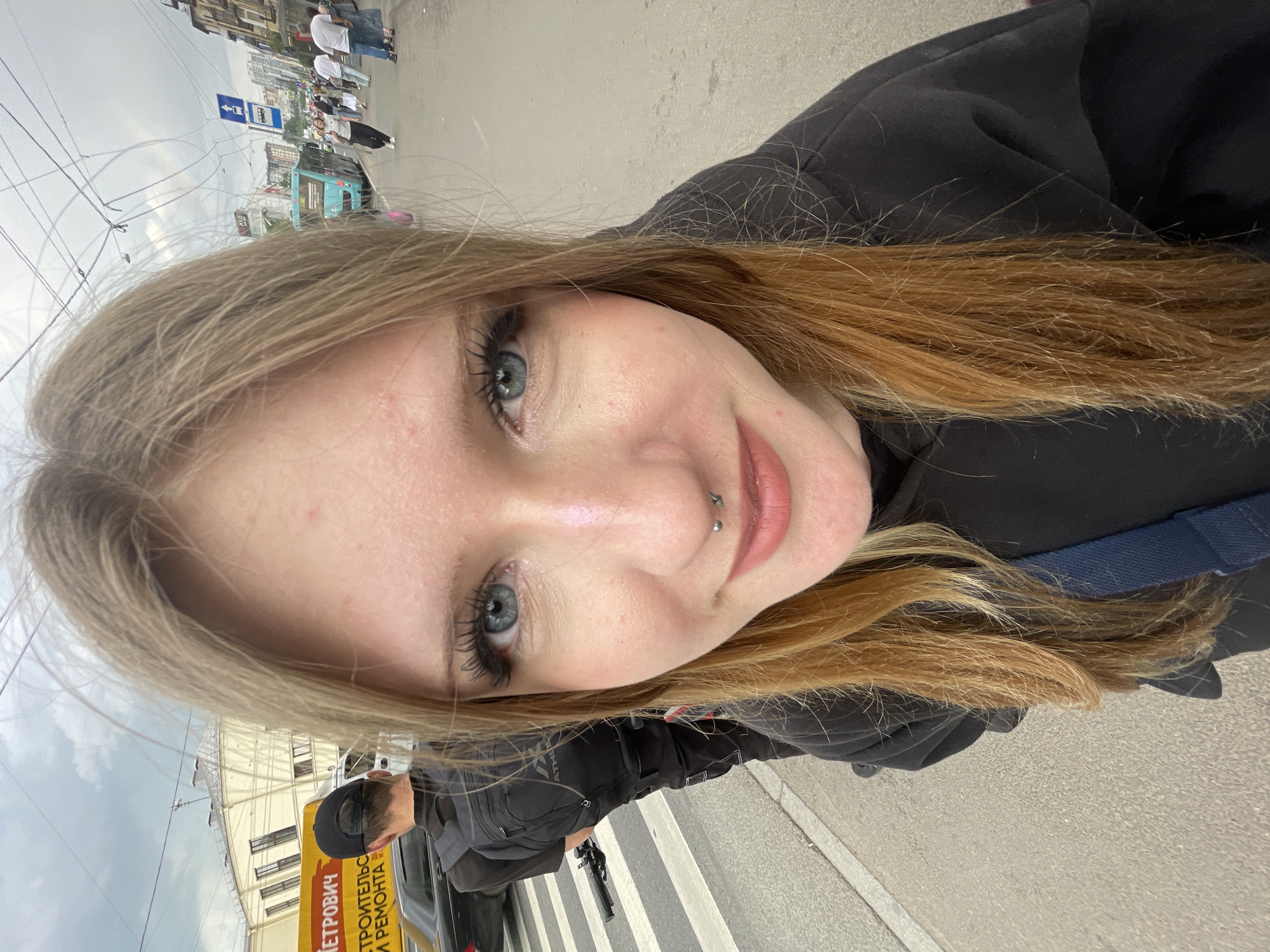 A young woman with light blonde hair, blue eyes, and a septum piercing takes a selfie in an urban setting. She has a faint smile and is wearing a dark top or jacket. The image is rotated 90 degrees clockwise, placing the subject's face in the foreground.
The background reveals a bustling city street in Saint Petersburg, Russia, under an overcast sky, suggesting daytime. Overhead wires, typical for trolleybuses or trams, are visible across the sky. Several blurred figures of people can be seen walking on a sidewalk, alongside buildings of varying architectural styles.
Key details include a partially visible yellow sign on the side of a vehicle, which displays Russian text: "...ПЕТРОВИЧЪ СТРОИТЕЛЬСТВО РЕМОНТА" (Petrovich Construction Repair). A blue street sign with an arrow is also visible further down the street. A pedestrian crossing with white stripes marks the road. The scene captures a casual moment amidst the urban environment.