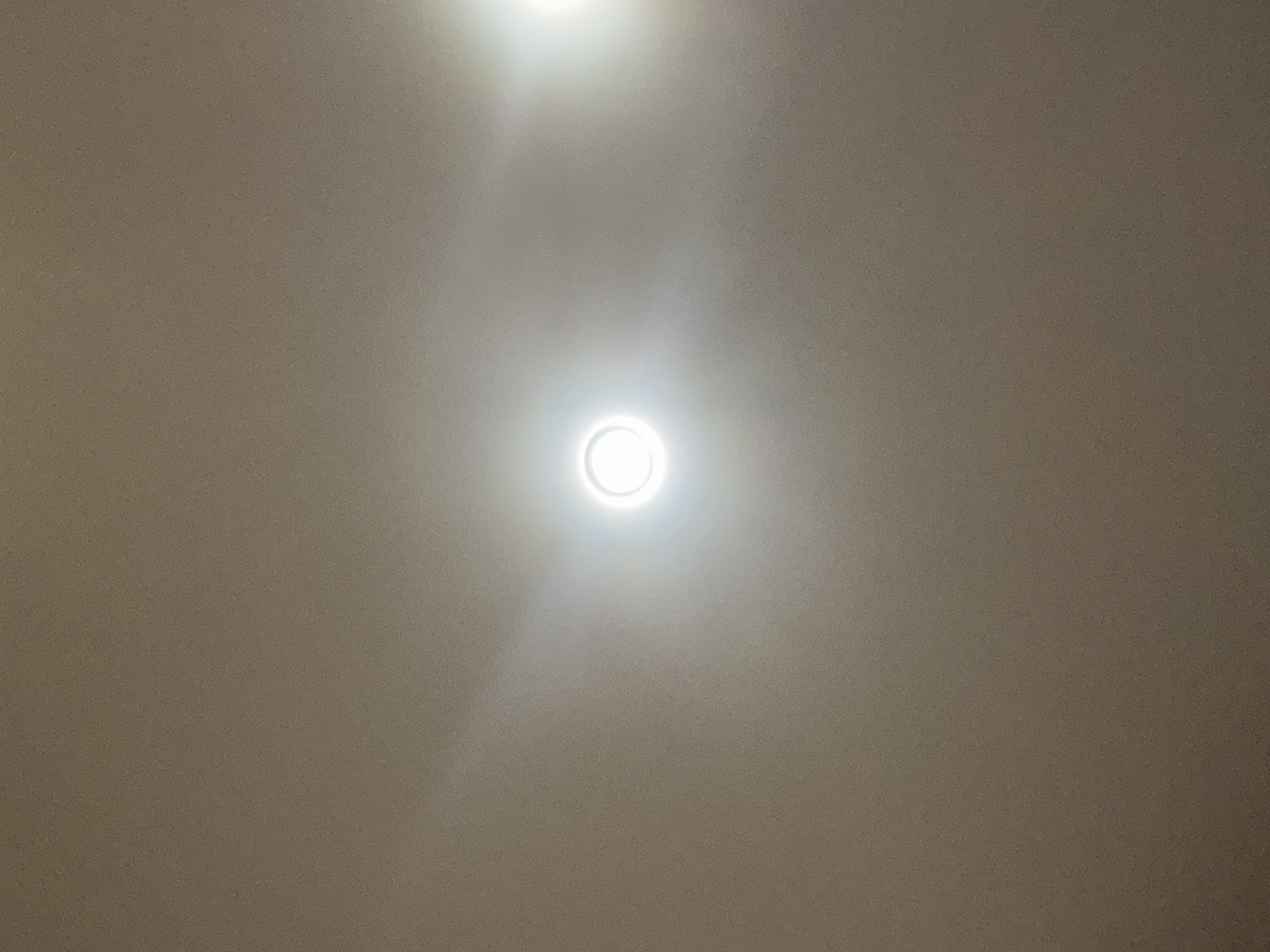 The image depicts a close-up, overhead view of a beige, textured ceiling. Two circular, recessed lights are visible. The light on the left is centered and emitting a bright white glow, with a distinct circular halo around it. The light on the right is positioned towards the upper right corner of the frame and is also emitting a bright white light, with a less pronounced halo. The texture of the ceiling appears somewhat grainy. There are no people, objects, or actions evident in the image, other than the illuminated ceiling fixtures. The context of Bugayevka, Russia, is not visually represented in the provided image.
