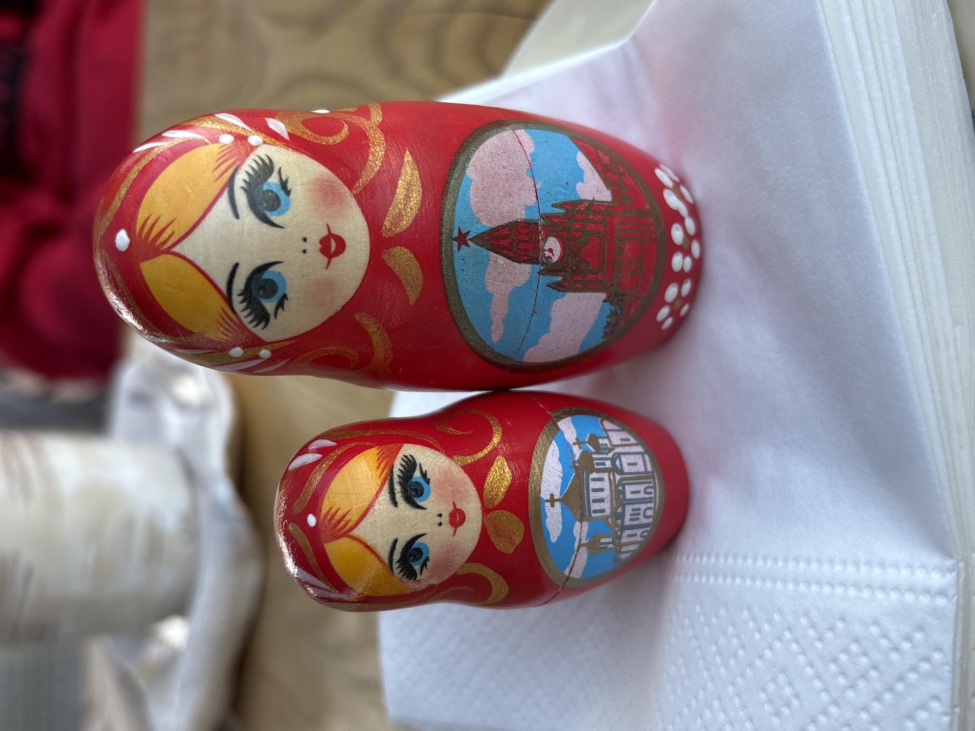 Two red Russian nesting dolls, known as Matryoshka dolls, rest on a white surface with a textured, possibly fabric-like appearance. The larger doll is positioned above the smaller one, both oriented in the same direction. Each doll is decorated with a traditional female face and depicts a different building scene within a circular frame. The top doll features an image of the Kremlin, specifically the Spasskaya Tower, in Moscow, Russia, identified by its distinctive red star. The lower doll displays an undefined building, possibly the State Regional Administration of Krasnoyarsk Krai, in Krasnoyarsk, Russia.