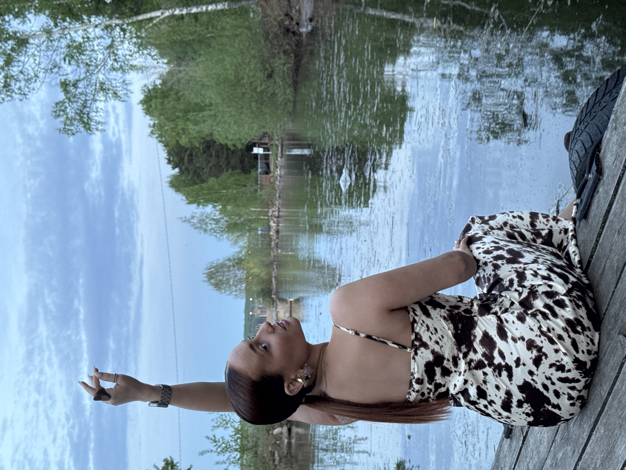 A woman is posing for a photo, likely on a wooden dock or pier next to a body of water. She is wearing a white and brown patterned dress, a watch, and jewelry. Her hair is pulled back. She is looking to her left and has one arm raised. The water reflects the sky and the trees along the shore. A tire can be seen on the dock near the woman. The location appears to be Chaikovsky, Russia, based on identifying elements of the landscape or urban setting.