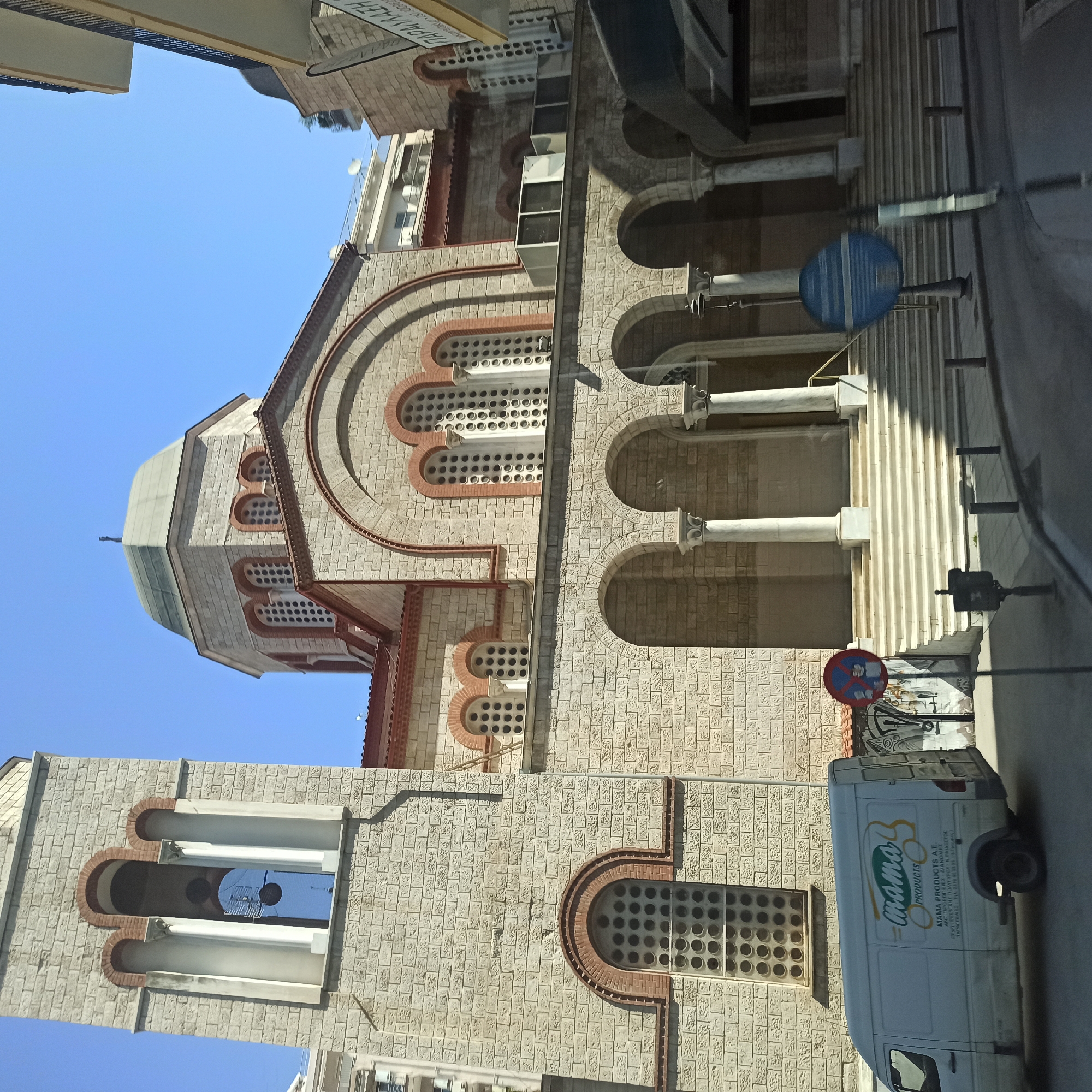 A van with "mama PRODUCTS" logo drives on a street in front of a stone church. The van is white with a logo that includes the word "mama" in a stylized script and the word "PRODUCTS" in a smaller font. The van is parked on the right side of the street. Behind the van, a church with a dome and a bell tower is visible. The church is made of stone and has arched windows. There are stairs leading up to the church entrance. In the background, there are buildings and a clear blue sky. The location is Thessaloniki Municipal Unit, Greece.