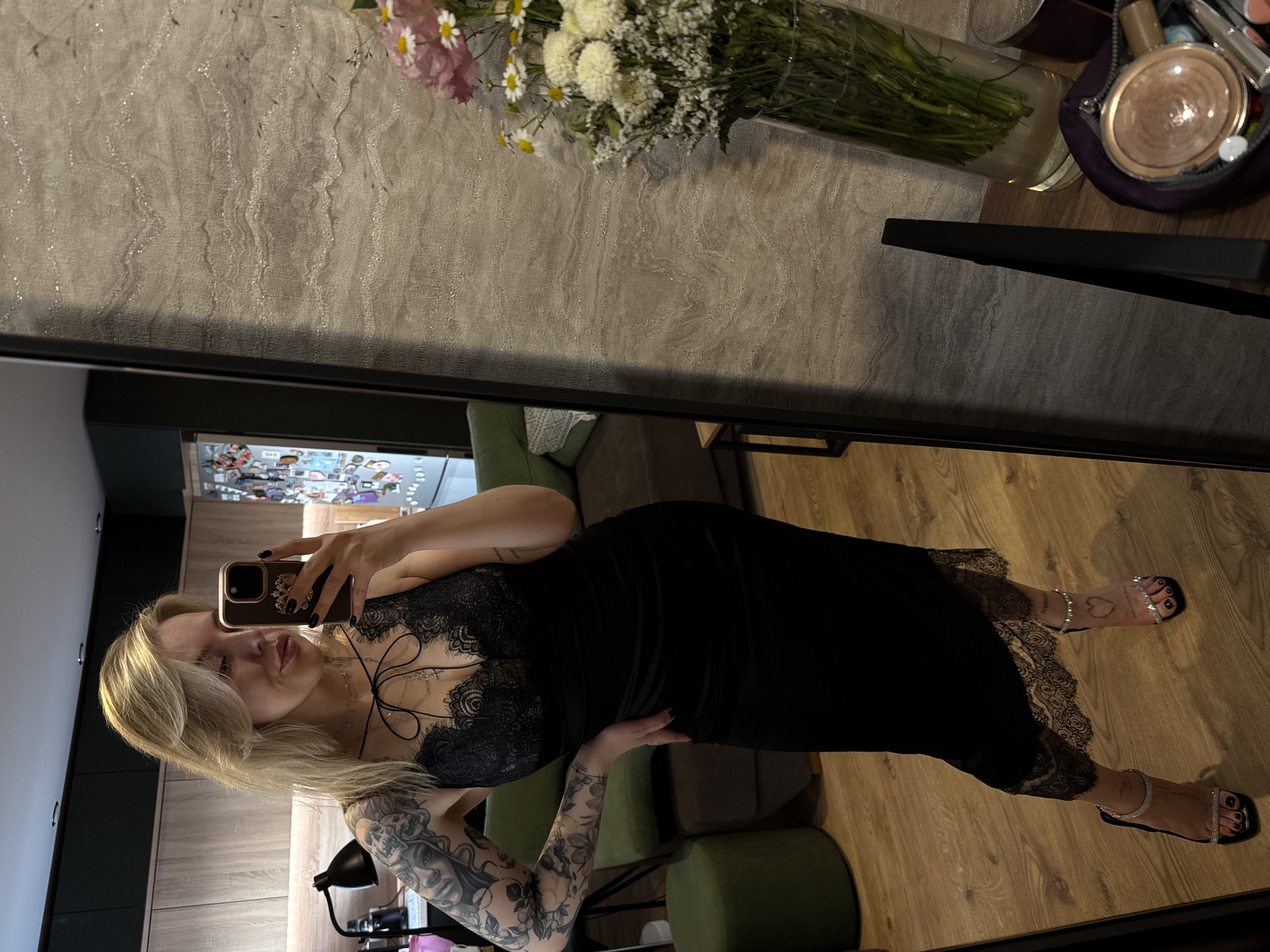 A woman with blonde hair and tattoos on her arms is taking a selfie in a mirror. She is wearing a black dress with lace detailing. Her feet are adorned with strappy sandals. The reflection in the mirror reveals a room with wooden floors and walls, as well as a display of decorative items and flowers. The location appears to be an interior space in Krasnoyarsk, Russia.
