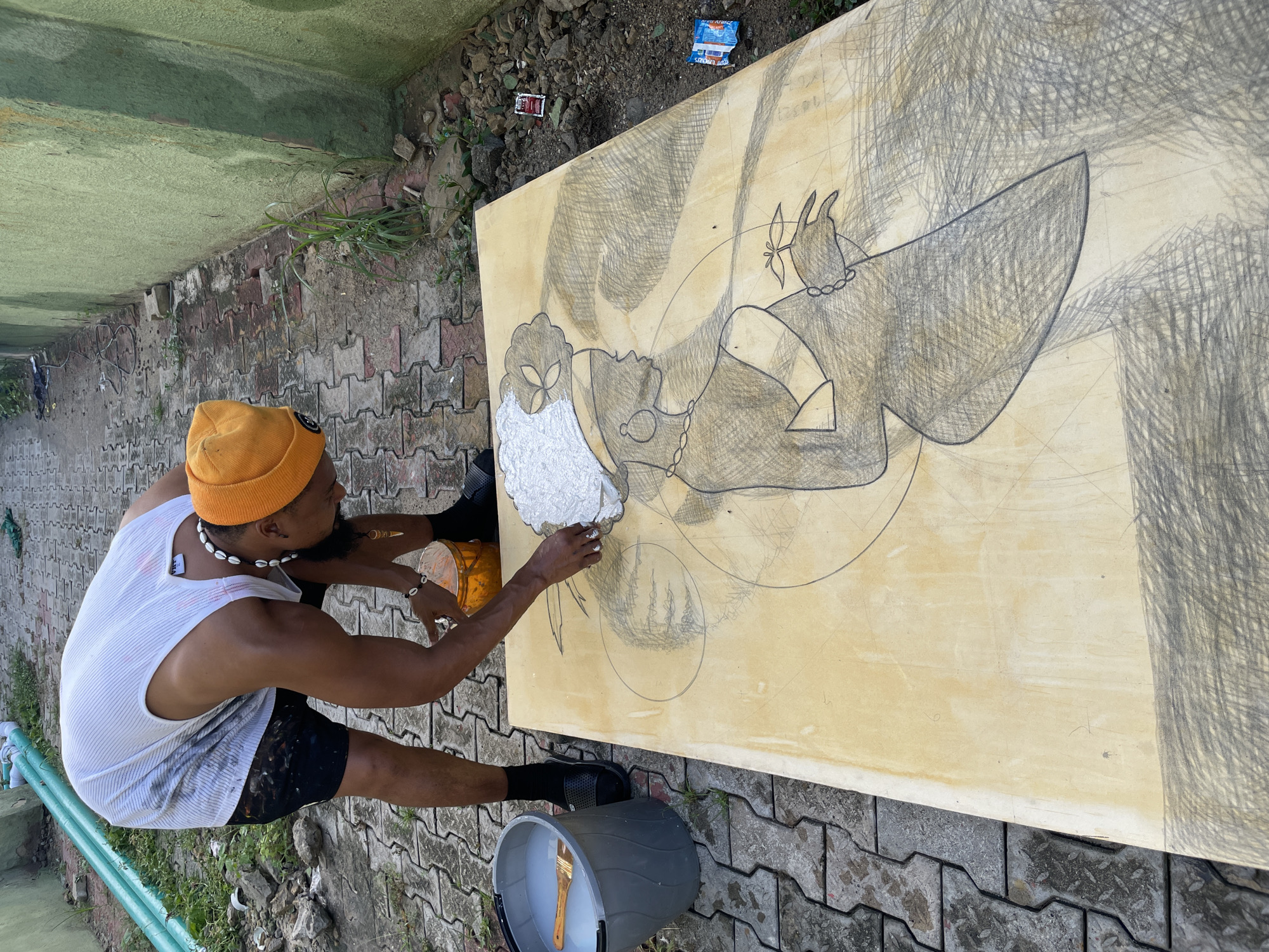 A man in Ado, Nigeria, is painting on a large canvas outdoors. He wears a yellow beanie, white tank top, black shorts, and shell jewelry. He is applying white paint to a portion of the canvas, which has a sketched figure in pencil. The canvas is on the ground, surrounded by a brick pavement and debris. A grey bucket with a paintbrush inside sits near the canvas. A small yellow container is visible near the artist's hands.