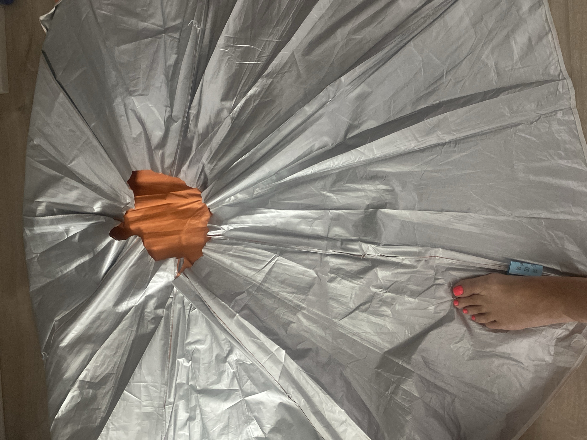 A person's foot, with bright pink toenail polish, is placed on a large, silver, tent-like object that is spread out on a light brown floor. The object is circular with pleats radiating from a central orange opening. The tent is likely made of a reflective material. A small blue tag with laundry care symbols is visible on the silver fabric near the foot. The scene appears to be indoors, possibly in a home in Novi Petrivtsi, Ukraine. The lighting is soft and even, suggesting indoor lighting or an overcast day. The overall impression is a domestic setting where the tent is either being set up, taken down, or stored.
