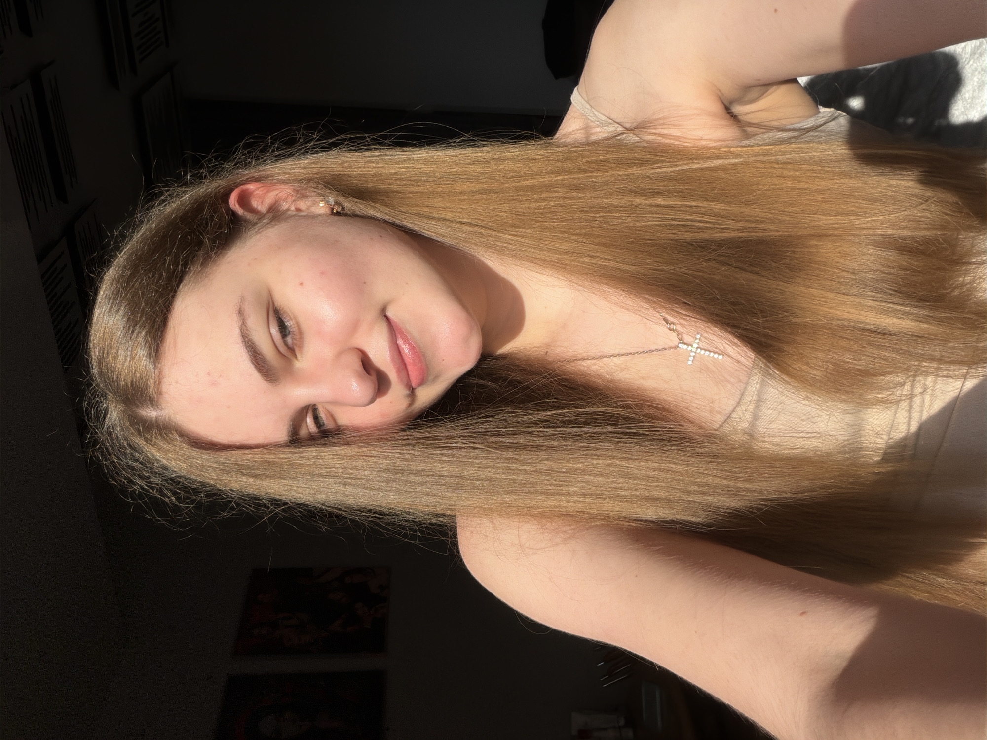 The image shows a young woman indoors, illuminated by natural light. She has long, straight, blonde hair and is wearing a light-colored camisole and a cross necklace. Her face is partially visible, with her head turned slightly towards the camera. The background is dark, with some framed pictures on the wall and what appears to be acoustic paneling on the ceiling. There are no discernible elements that would indicate the location is in Dnipro, Ukraine, or any other specific city. The scene appears to be a personal photograph taken in a home environment.