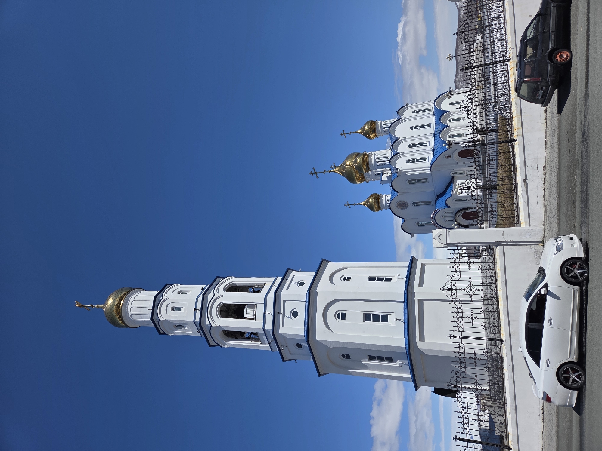 The image depicts a white and blue Orthodox church complex with golden domes, under a clear blue sky. A tall bell tower is prominently featured on the left, while the main church building is on the right, both enclosed by a decorative iron fence. A white car is parked on the right side of the church, and further beyond, a dark-colored car is visible. The location appears to be urban, with a paved road adjacent to the church. The city is Unknown, Unknown.