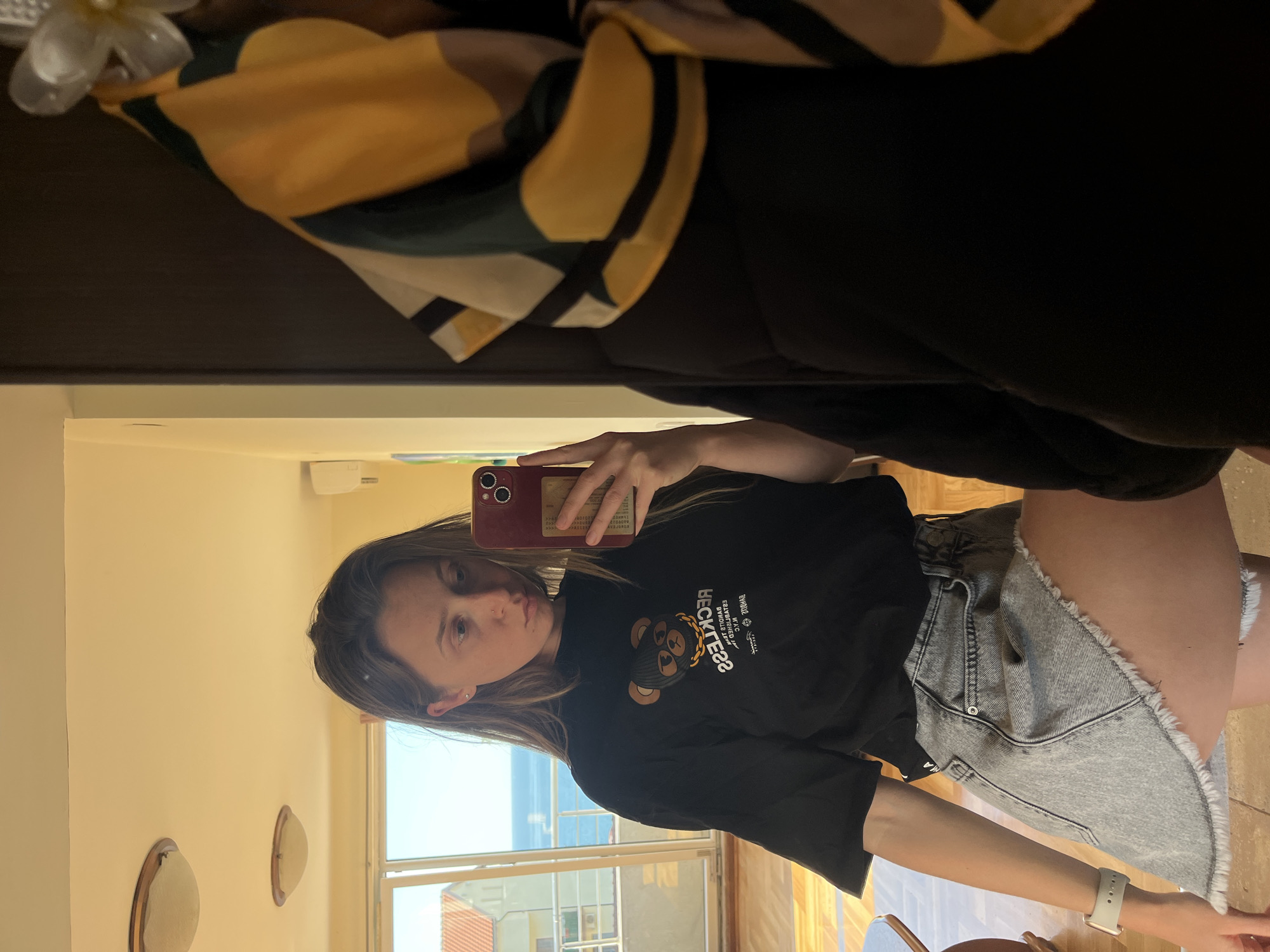 A woman is taking a mirror selfie in a room with a window overlooking Bečići, Montenegro. The room has beige walls with two oval decorative wall pieces. Outside the window, one can see buildings with red tile roofs and the Adriatic Sea. The woman is wearing a black T-shirt with a cartoon bear graphic and frayed denim shorts. She is holding a red phone. A piece of patterned yellow and green fabric is visible hanging above the mirror.