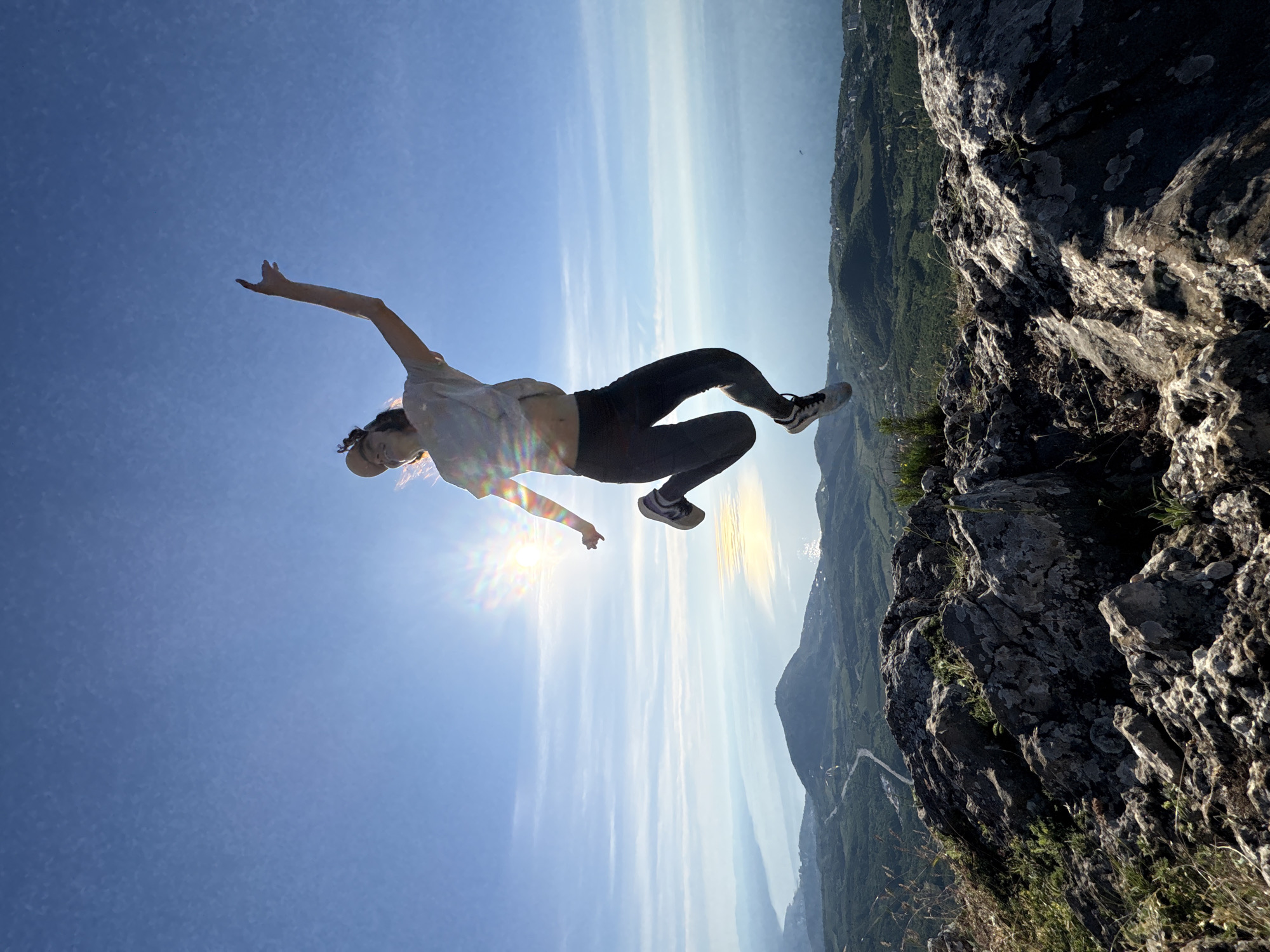 An adult individual is captured mid-air, jumping on a rocky mountain peak overlooking a vast coastal landscape under a bright blue sky. The location is an unnamed mountain in Unknown, Unknown. The person, appearing female, is silhouetted against the sun, which is positioned to her upper left, emitting a strong lens flare and starburst effect. She is wearing a light-colored t-shirt, dark leggings, and athletic shoes, along with a light-colored baseball cap. Her right arm is raised with the hand open, fingers splayed upwards, while her left arm is bent close to her body. Her legs are bent, indicating the peak of a jump.
The foreground consists of rugged, greyish-brown rocks with visible textures, lichen, and scattered patches of green grass and small plants. Below the immediate peak, the midground reveals a sprawling green landscape of rolling hills covered in dense vegetation, with a winding road traversing through it. Another prominent mountain or large hill is visible further back in the midground. In the far background, a large body of water, likely a sea or ocean, stretches to the horizon, reflecting the sunlight in a shimmering strip. Distant landmasses are faintly visible across the water. The sky is clear and bright blue with minimal cloud cover. The overall scene conveys a sense of exhilaration and vastness from a high vantage point.