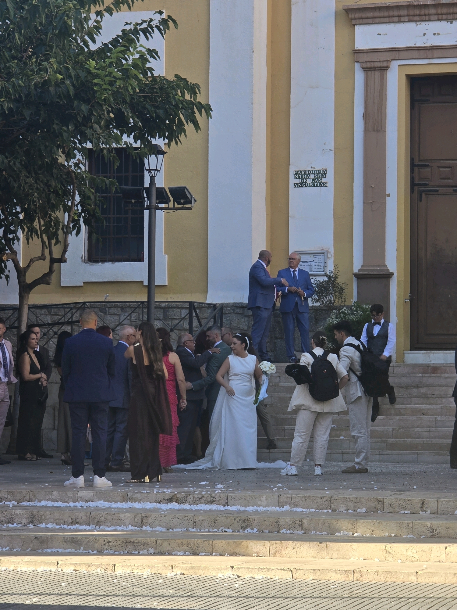 The image captures a gathering outside a building, likely a church, given the architectural style and the presence of a wedding party. The building is painted in alternating bands of yellow and white, with brown architectural details around a large wooden door on the right. A sign on the wall reads "PARROQUIA NTRA SRA DE LAS ANGUISTIAS." The foreground shows a set of stone steps leading up to the entrance, scattered with white confetti.
Several individuals are visible in the scene, dressed in formal attire. On the steps to the left, a bride in a white wedding dress is holding a bouquet. Nearby, several guests are seen, including women in long dresses and men in suits. Two men in matching blue suits are engaged in conversation on the steps, appearing to be officiators or guests of importance. Further to the right, a group of younger individuals, some wearing backpacks, are also on the steps. The overall atmosphere suggests a wedding ceremony or celebration in progress. A tree with dark green foliage is present on the left side of the image, providing shade. The lighting indicates it is daytime. The location is identified as the Parish of Our Lady of Anguish in Málaga, Spain.