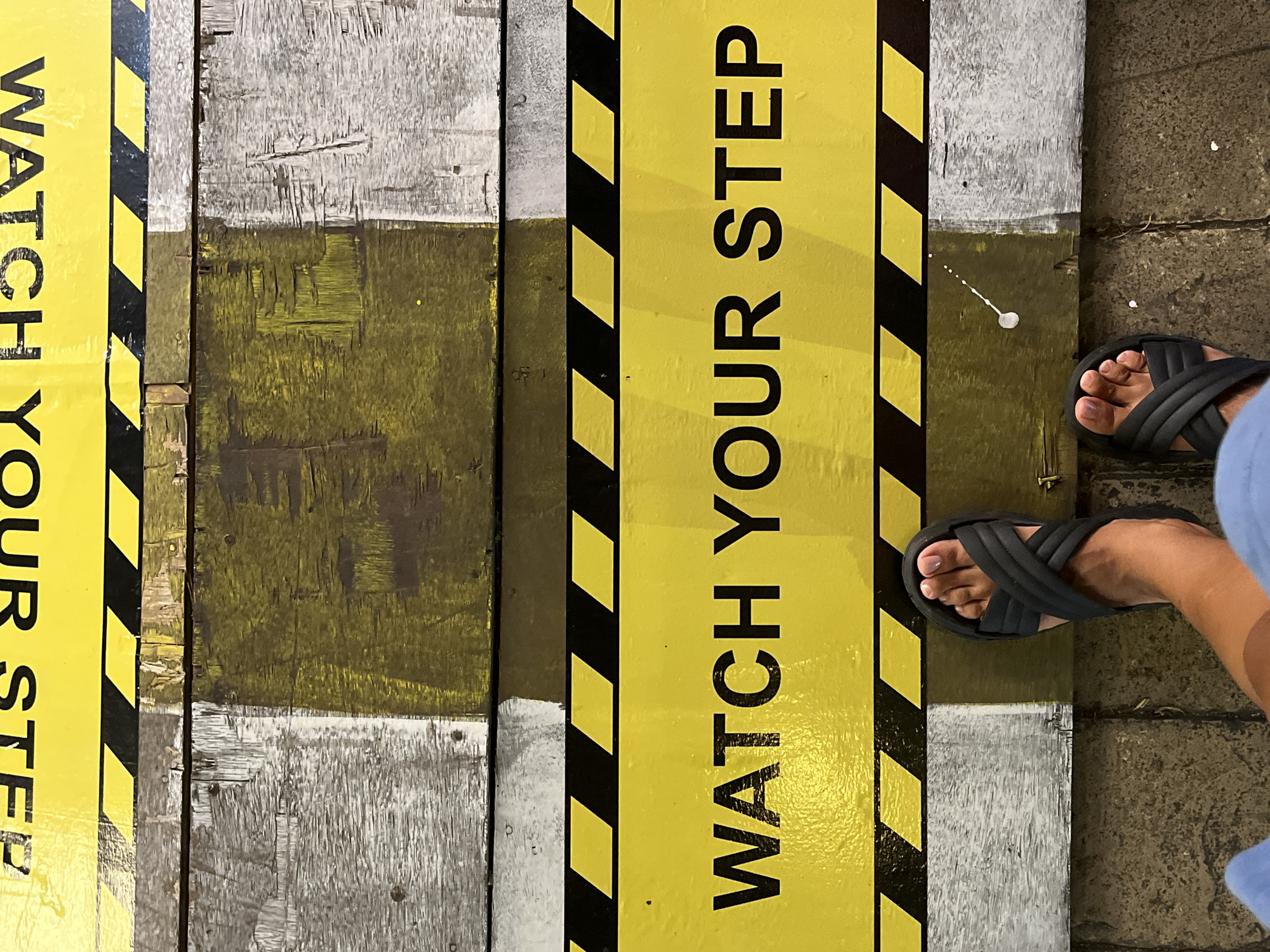 The image shows a person's feet in black sandals standing on a wooden walkway with the words "WATCH YOUR STEP" painted in black on a yellow background. The walkway is constructed of weathered planks, some painted yellow and some gray. Black and yellow caution stripes border the yellow section. The location is unknown, as no indicators of Kuta Selatan, Indonesia are visible.
