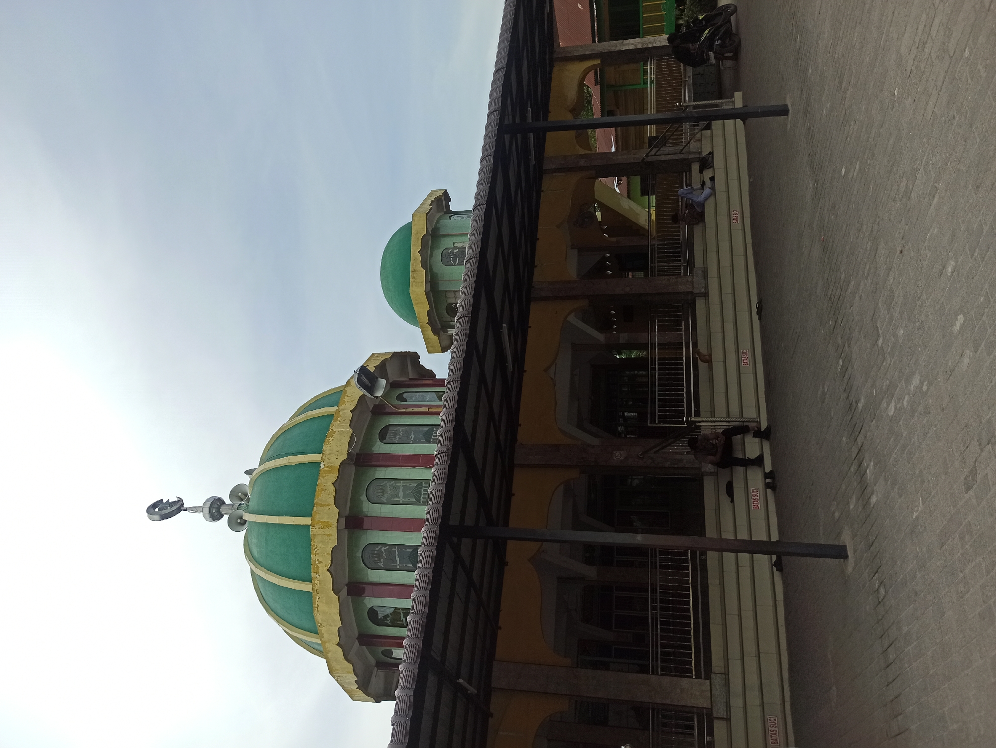 This is a full-frame, eye-level outdoor shot of a mosque in Medan, Indonesia. The mosque features a large green dome with a crescent moon and star on top, and a smaller green dome. The building has yellow and maroon accents, with arched windows. A covered walkway extends in front of the building, supported by columns. Several people are visible sitting on the steps leading up to the mosque, and a motorcycle is parked to the right. The ground is paved with interlocking bricks. The sky is overcast and pale. The overall scene suggests a public religious space in a tropical climate.