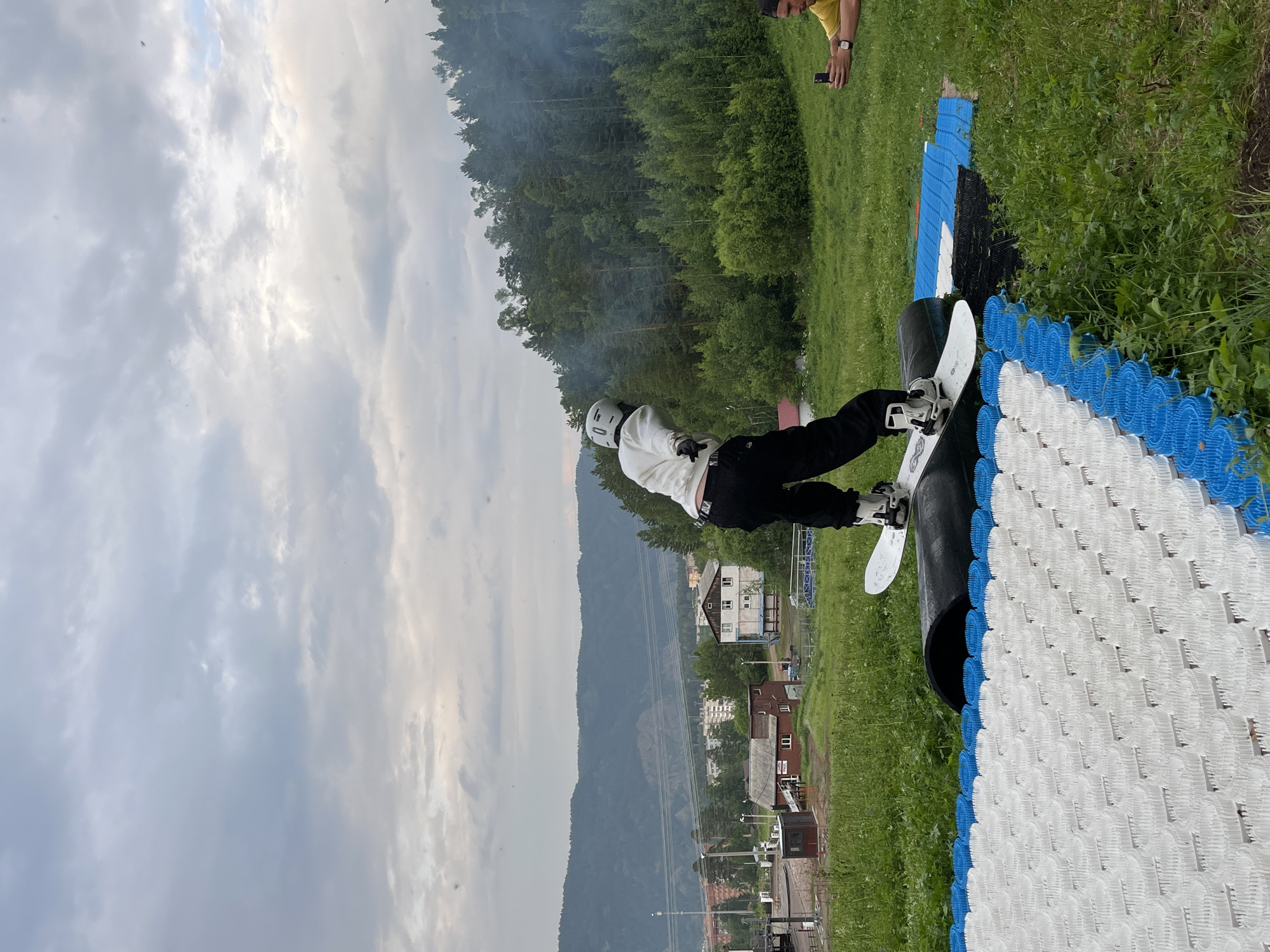 A person wearing a white helmet, white long-sleeved shirt, and black pants is snowboarding down a black cylindrical obstacle. The snowboarder is positioned mid-action, with their left foot forward on the snowboard. They are riding over a textured surface made of blue and white interlocking pieces. To the right of the snowboarder, another person in a yellow shirt is holding a smartphone, seemingly filming or taking pictures. The background depicts a lush green grassy hill leading up to a dense forest. In the distance, there are buildings and mountains, suggesting a town or city on the outskirts of a natural landscape. The sky is overcast with grey clouds. This scene is likely at a recreational area or training facility, possibly associated with outdoor sports, in or near Divnogorsk, Russia, given the mountainous terrain and visible infrastructure.