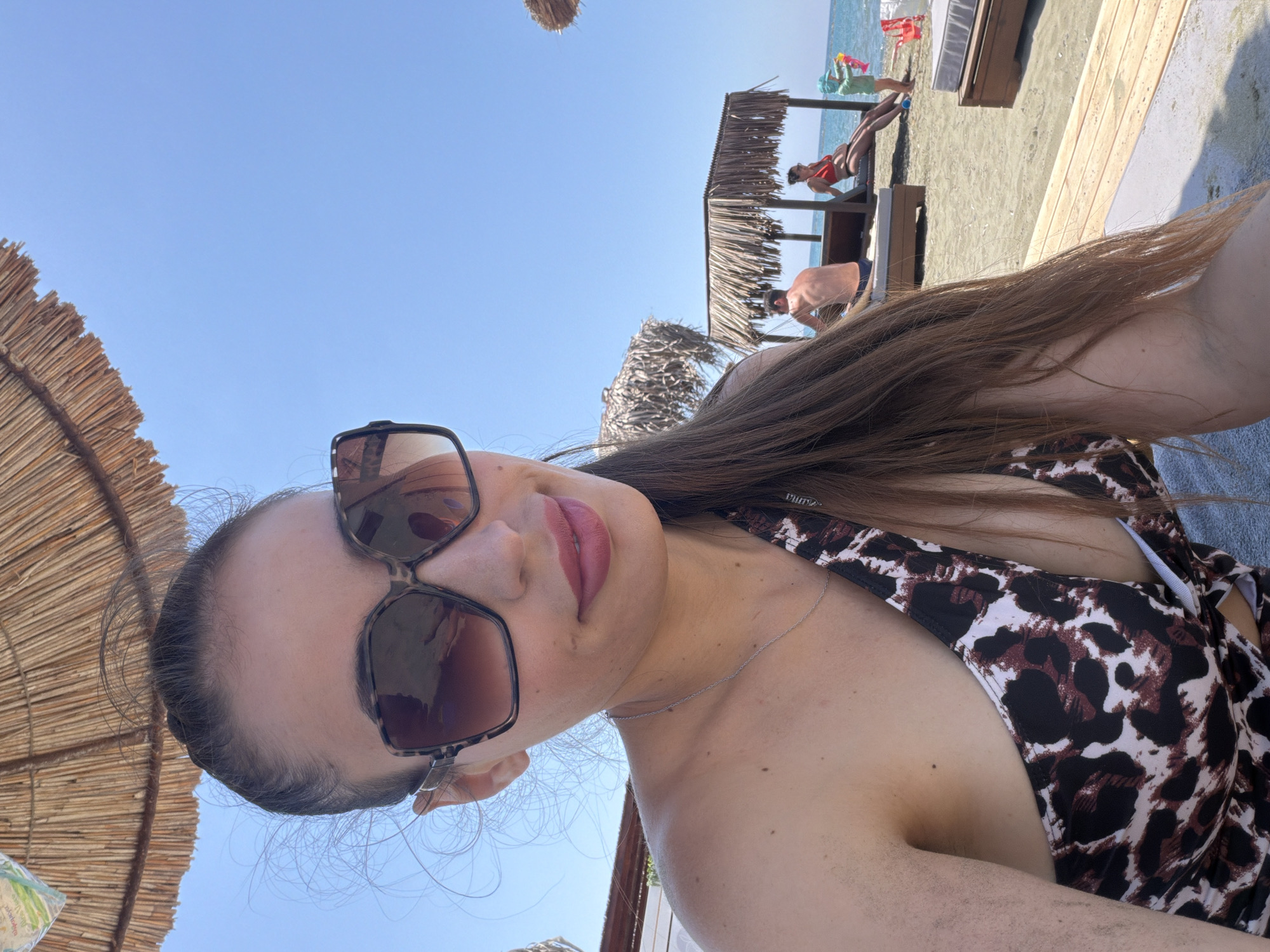 This image depicts a sunny beach scene in Meneou, Cyprus. In the foreground, a woman, appearing to be in her late 20s or 30s, is captured from a slightly low angle, likely in a selfie pose. She wears large, dark-tinted sunglasses with a tortoise-shell frame, dark pink lipstick, and a leopard-print swimsuit featuring black, brown, and cream patterns. Her long, dark brown hair is styled back, with a few strands loose around her face. A thin silver chain necklace is visible on her chest. The background reveals a sandy beach extending to a clear blue ocean under a bright, cloudless sky. Several thatched sun umbrellas and a larger, square-shaped thatched cabana are positioned on the sand. Under the cabana, a shirtless man in dark swim shorts sits on a lounge chair with his back to the camera, while a woman in a red swimsuit sits on an adjacent lounge chair, facing the water. To the right of the cabana, a young child wearing a light blue hat plays at the water's edge with red and yellow beach toys. Other lounge chairs are visible on the beach.