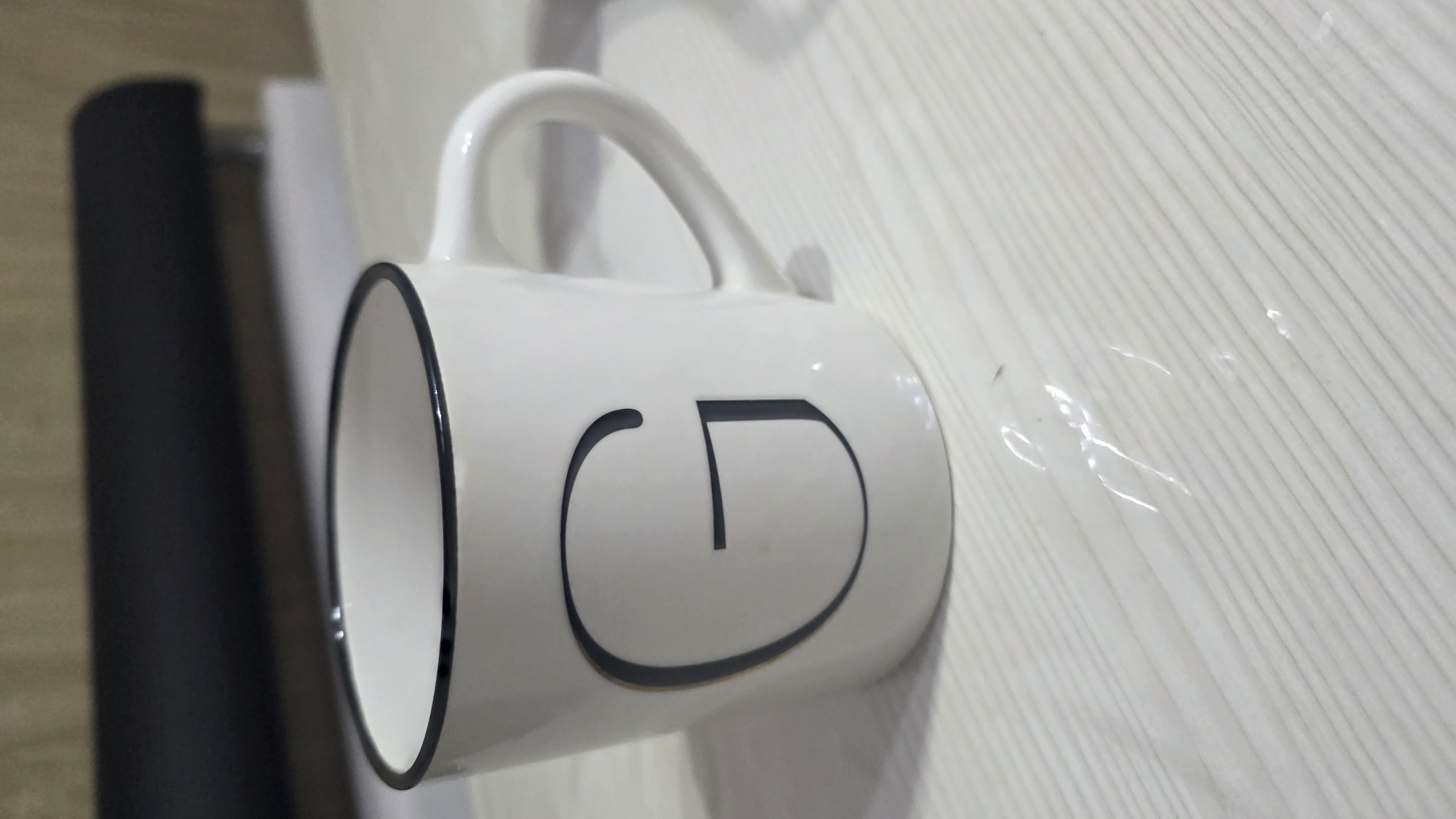 The image shows a white ceramic mug with a black rim and a large black letter "G" printed on its side. The mug is placed on a white, horizontally-lined surface that appears to be a table. In the background, a blurred dark object, possibly a stapler, is visible on top of a white surface. The lighting is soft and diffused, casting subtle shadows. There are no people or other objects of significance in the frame. The scene is static and does not depict any activity or interaction. The location in the city of Chuy, Kyrgyzstan, is not visually indicated in the image.