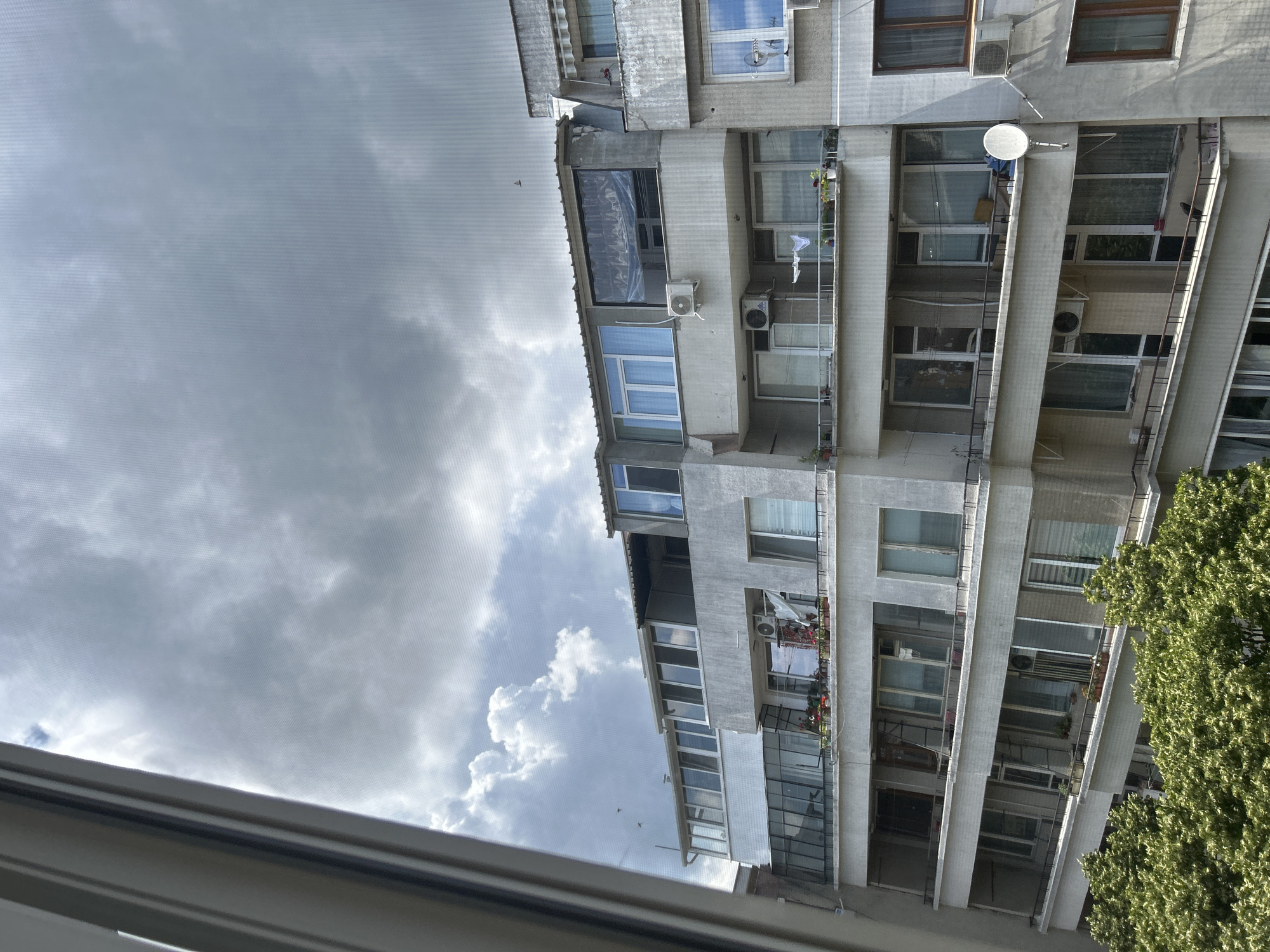 The image shows a building in Varna, Bulgaria, with multiple floors and balconies. The sky is cloudy with some patches of sunlight. The building appears to be residential, with several windows and air conditioning units visible. There are trees in the foreground, suggesting a green area or park nearby. The overall scene is of a typical urban environment in Varna.