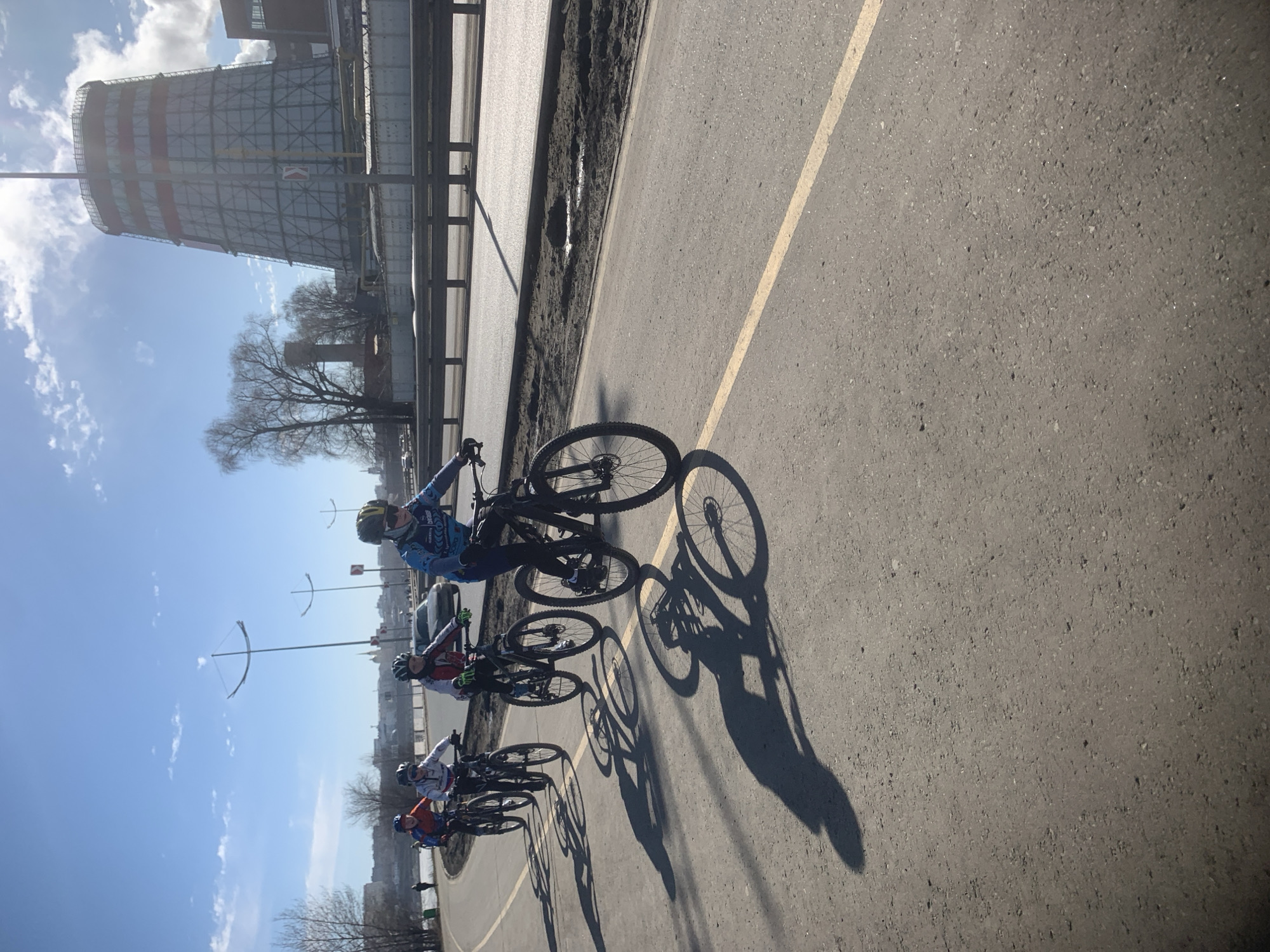 The image depicts a group of cyclists riding on a paved road alongside a river in Izhevsk, Russia. There are four cyclists visible, each wearing cycling gear and helmets. The road has a yellow lane marker. Adjacent to the road is a metal railing and a patch of land. In the background, a large industrial structure, possibly a cooling tower, dominates the skyline, along with bare trees under a partly cloudy sky.