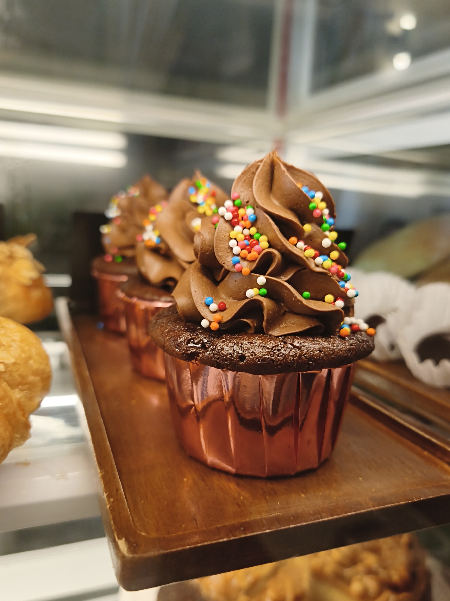A close-up, eye-level shot showcases a row of three chocolate cupcakes adorned with swirled chocolate frosting and colorful sprinkles. The cupcakes are presented in shiny, rose-gold foil wrappers and rest on a wooden tray. In the background, other baked goods, including croissants, are visible within a display case. The lighting suggests an indoor setting, likely a bakery or cafe. The scene is devoid of people, with the focus solely on the confectionery items. The overall impression is one of freshness and appealing presentation.