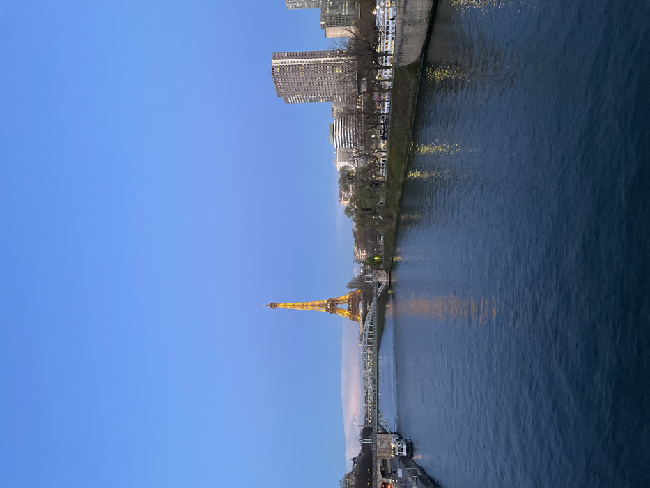 The image presents a wide view of the River Seine in Paris, France, at dusk, looking eastward towards the Eiffel Tower. The sky above is a deep blue, transitioning to a lighter, faint pinkish hue near the horizon on the left.
The central and most prominent subject is the Eiffel Tower, situated in the middle distance, fully illuminated in a warm, golden light. Its illumination is reflected on the surface of the dark blue water of the River Seine, which occupies the foreground and midground of the scene, flowing from right to left. The water is textured with ripples.
A multi-level bridge, identified as the Pont de Bir-Hakeim, spans the river in the midground, positioned between the viewpoint and the Eiffel Tower. Its structural elements are visible, and the upper level appears to carry a roadway while the lower level accommodates a metro line.
Along the riverbanks, bare trees line the promenades, suggesting a colder season. On the left (south) bank, an urban structure or entrance is visible near the water, with at least two docked boats or barges. On the right (north) bank, a series of streetlights illuminate the promenade, behind which various urban buildings are visible, including a tall, multi-story residential or office block and several other white, angular structures. Several illuminated passenger boats are moored along the right embankment. No individual people are distinctly visible within the frame.