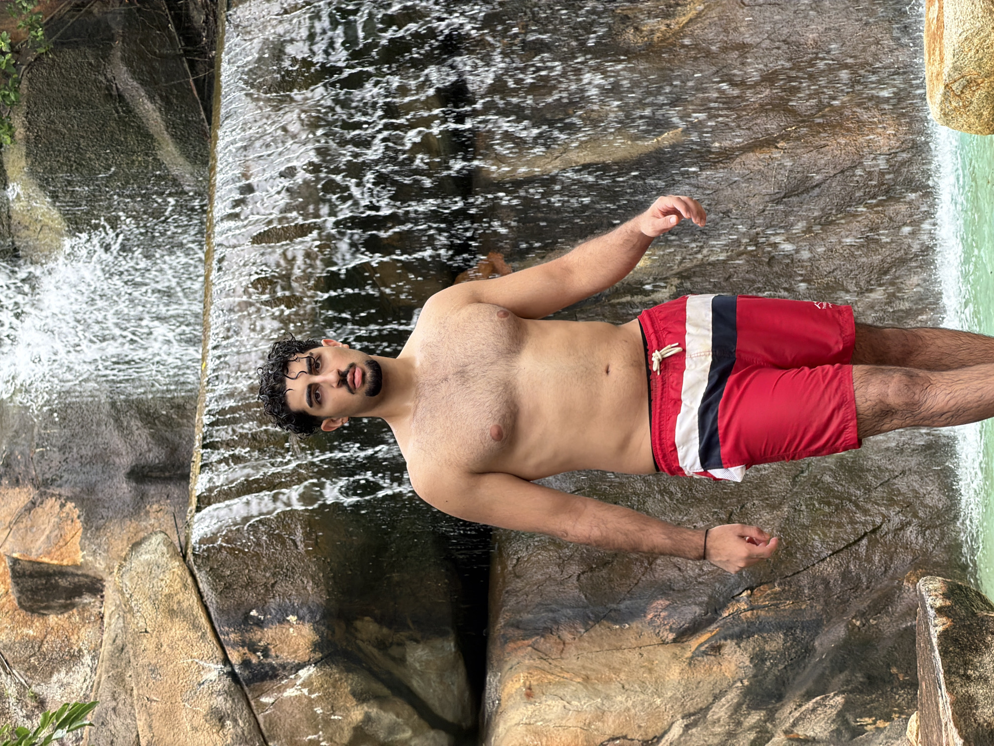 A young man with dark curly hair and a beard stands in front of a waterfall, wearing red swim trunks. He is shirtless, revealing his chest and arms. The waterfall cascades down a rocky cliff face, with water splashing into a turquoise pool below. The scene is set in a natural outdoor environment, likely a park or nature reserve, given the presence of rocks and foliage. The man appears relaxed and is looking directly at the camera. The location is identified as Nha Trang, Vietnam.