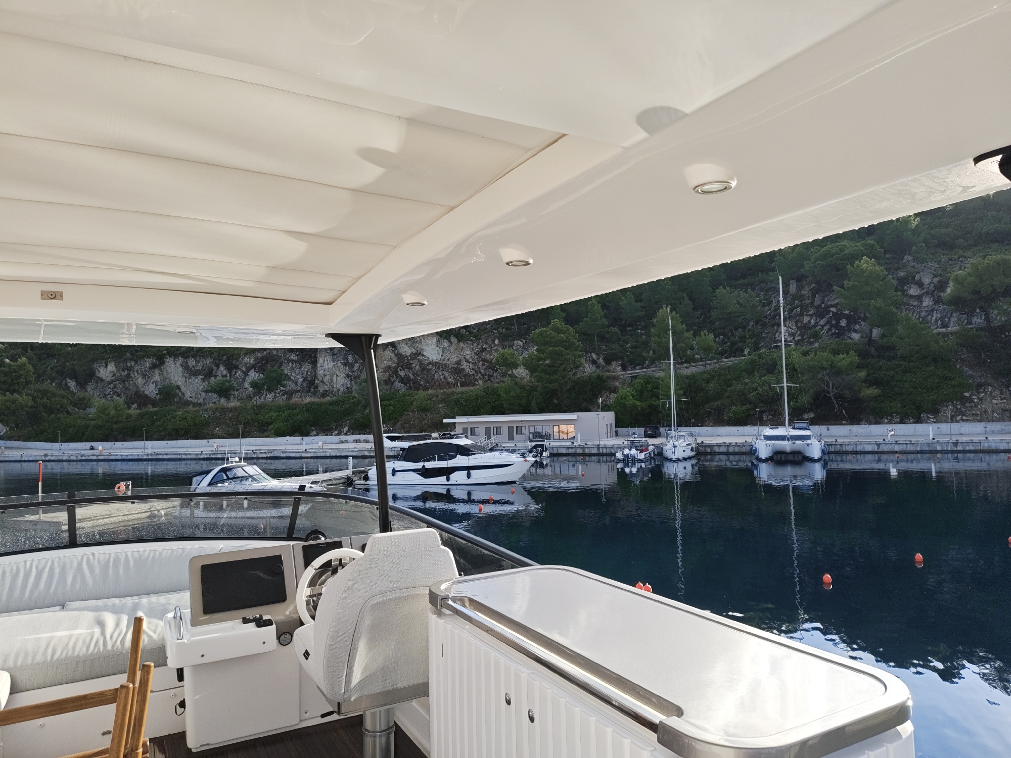 The image is an exterior, eye-level shot from a white yacht docked in a marina. The yacht has a white canopy overhead with recessed lighting. In the foreground, a white leather seat and a portion of the yacht's helm with a navigation screen are visible. To the left, a wooden ladder and white seating are present. The marina is calm and reflects the surrounding environment. Across the water, several other boats are docked, including a white motor yacht and sailboats. A stone embankment lined with trees forms the backdrop, with a building visible in the mid-ground. The scene is set during the day, with the sun casting shadows and reflecting off the water. The location is identified as Sithonia Municipal Unit, Greece.