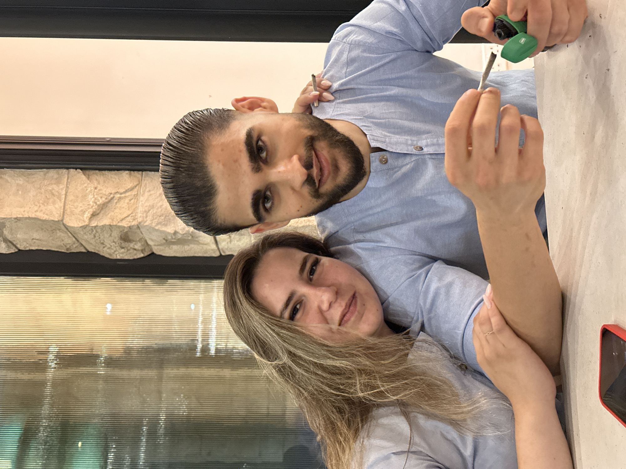 A light blue-shirted person with light brown hair rests their head on the shoulder of a person in a light blue shirt. The person with light brown hair is looking at the camera with a slight smile. The person in the light blue shirt has dark hair and a beard, and is looking at the camera with a smile. Their hand is holding a cigarette lighter and a cigarette. They are sitting at a table in a restaurant. The background shows a glass partition with vertical lines and a stone wall. This is likely a restaurant or cafe in Nicosia, Cyprus.