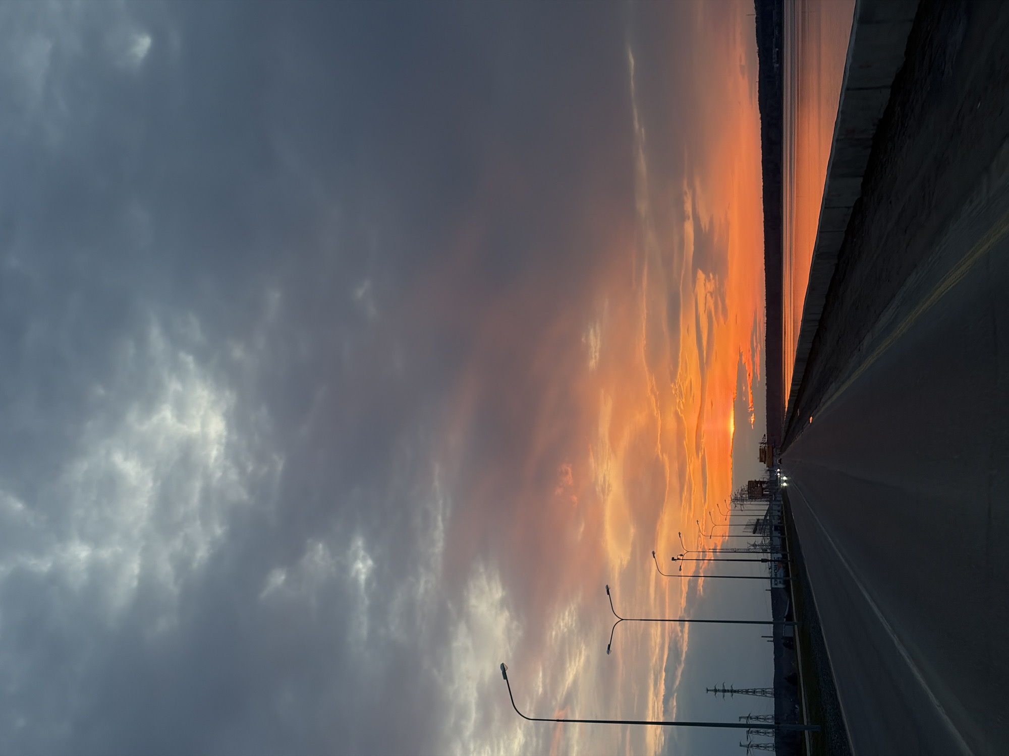 The image captures a sunset in what appears to be the city of Chaikovsky, Russia. A wide asphalt road stretches into the distance, flanked by a concrete barrier on the right and a row of evenly spaced streetlights on the left. Beyond the barrier is a body of water reflecting the colors of the sunset. Overhead power lines and support structures are visible on the left side of the road. The sky is a mix of dark, gray clouds and streaks of orange, pink, and yellow from the setting sun. The sun itself appears to be on the horizon, obscured by the clouds, with intense orange light radiating around it.