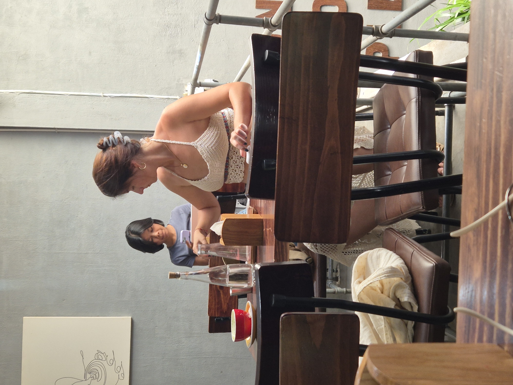 The image captures a moment at a cafe in Đà Nẵng, Vietnam. Two women are seated at a wooden table. In the foreground, a woman with brown hair tied up in a bun is wearing a white crocheted top and a necklace. She is looking down and interacting with something on the table. To her left, a bottle of water and a red cup are visible on the table. Behind her, another woman with short black hair is seated and looking at her phone. The cafe appears to have a minimalist aesthetic with grey walls and wooden furniture. There are also metal railings and some greenery visible in the background. The overall lighting suggests it is daytime.