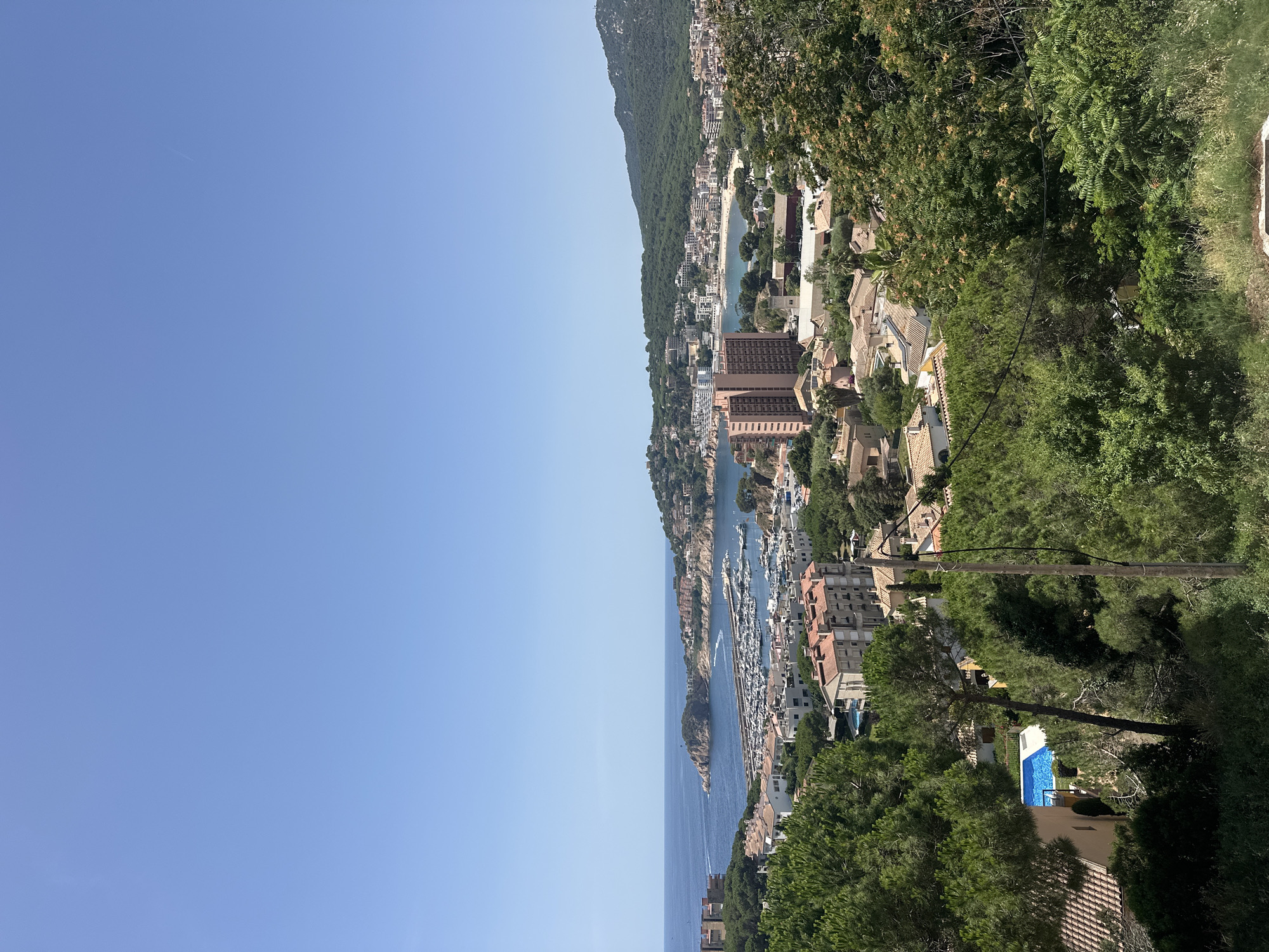 This is a high-angle, medium shot of a coastal town in Sant Feliu De Guíxols, Spain, under a clear blue sky. In the foreground, dense green trees and foliage partially obscure the view of terracotta-roofed buildings and a bright blue swimming pool. The middle ground reveals a marina filled with numerous white sailboats and yachts, situated within a bay. Beyond the marina, the town sprawls along the coastline, with a mix of low-rise residential buildings and a prominent tall, salmon-colored apartment building. Further in the distance, a lush, green hillside forms the backdrop, leading to a mountain range under the expansive sky. There are no people or animals visible in the scene. The overall impression is of a sunny, Mediterranean coastal resort.