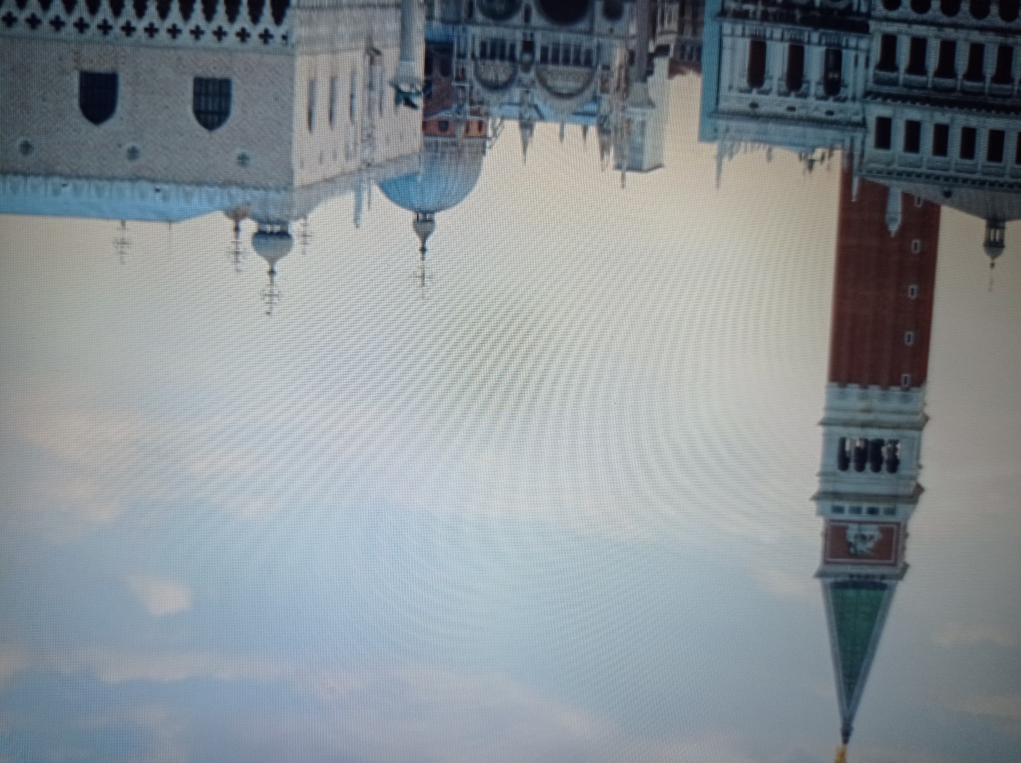 The image shows a cityscape with several prominent buildings and a tall tower. The architecture suggests a European city, possibly Venice, Italy, with its characteristic Venetian Gothic style. The foreground features detailed facades of buildings, some with arched windows and decorative elements. In the background, a tall, red brick tower with a green pyramidal roof dominates the skyline. A large dome-shaped structure is also visible, further suggesting a place of historical and architectural significance. The sky is a pale blue with some scattered clouds. There are no people visible in the image. The question states the location is Mukachevo, Ukraine, however, the architectural style of the buildings strongly suggests Venice, Italy, rather than Mukachevo.