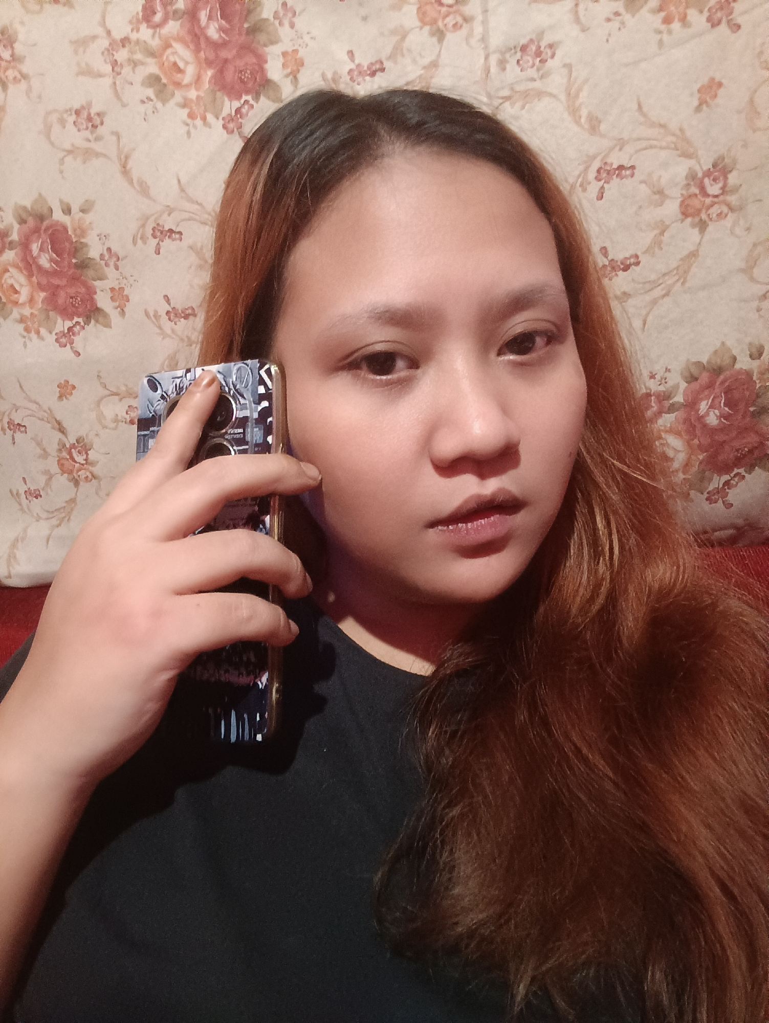 A young woman with long, auburn hair is holding a smartphone to her right ear, obscuring part of her face. She is wearing a black t-shirt and is positioned in front of a floral-patterned wallpaper. The lighting is dim, and the image appears to be taken indoors. The woman's left hand is visible, holding the phone. The backdrop is a wall with a repeating pattern of large, pink and red roses with green leaves and smaller pink flowers. The scene is likely captured in Depok, Indonesia, given the context provided, though no specific identifying landmarks are visible.