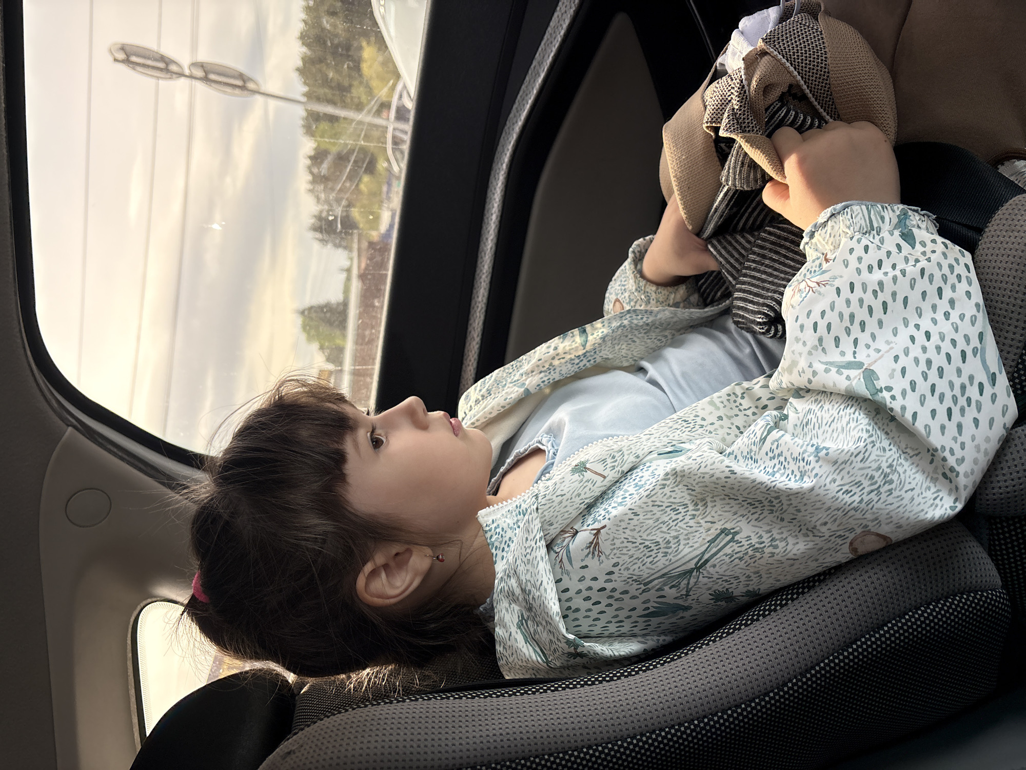 A young girl is sitting in a car seat. She has brown hair and bangs, and is wearing a patterned jacket. She is holding a pile of clothes. The car is moving and there is a view out the window, including a street light. The location is potentially near Chaikovsky, Russia, as indicated by the street light design that is found within that region.