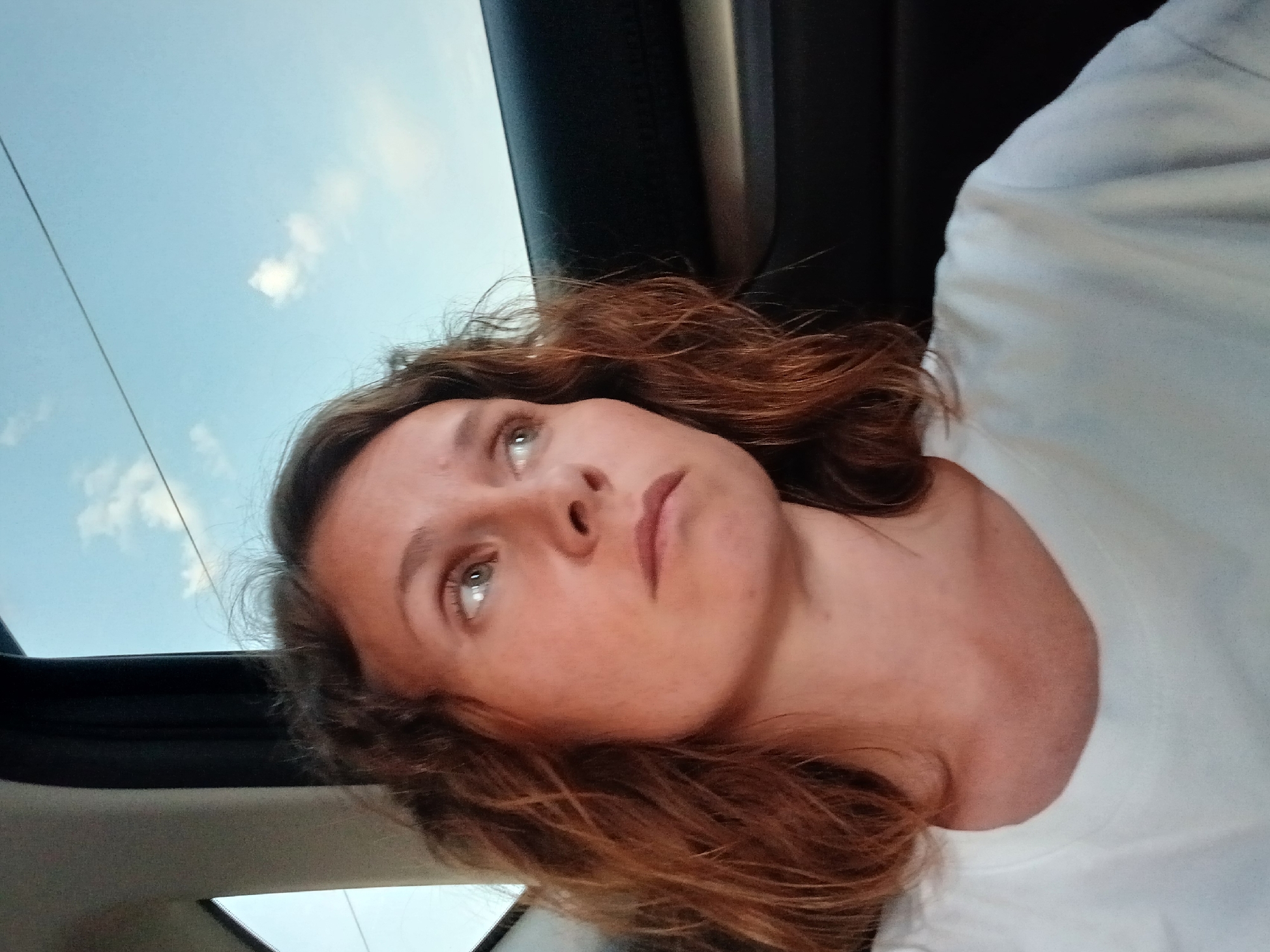 A woman is seen from the chest up, her head turned to her right. She is wearing a white t-shirt and has shoulder-length, wavy brown hair. The background shows a light blue sky with a few white clouds and what appears to be a power line. The setting seems to be inside a vehicle, with part of a car's interior visible on the right side of the image. The woman is looking out the window, her expression thoughtful. The lighting suggests it might be late afternoon or early evening. The image was captured in the city of Yalgyz-Narat, Russia.