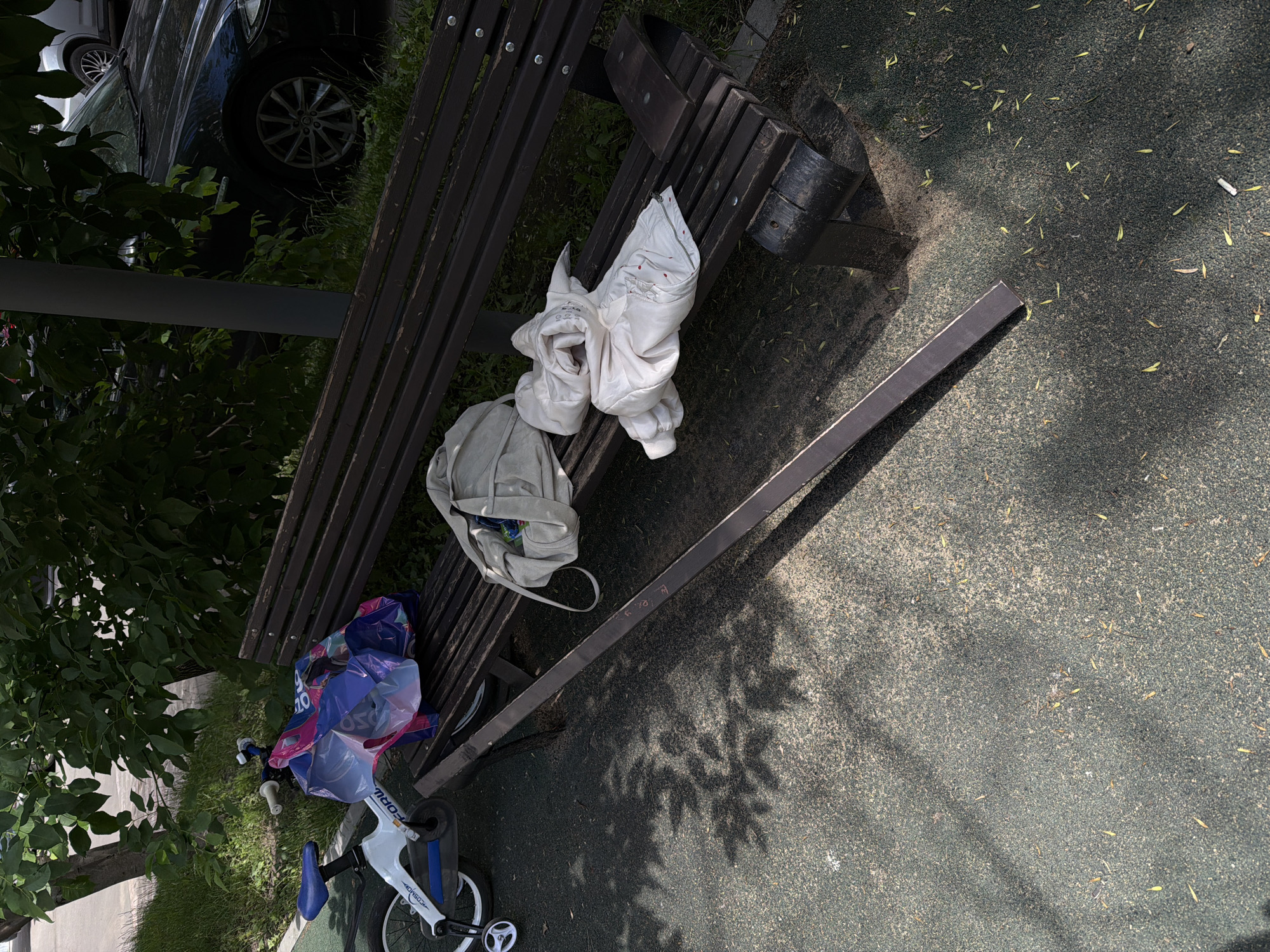A bicycle is parked next to a wooden park bench. Several bags and a white jacket are placed on the bench. The scene appears to be in a park or public space, possibly in Moscow, Russia, given the presence of trees and a dark-colored car in the background. The ground is covered with a dark, speckled surface, possibly rubberized playground material, with scattered leaves and shadows cast by the trees.