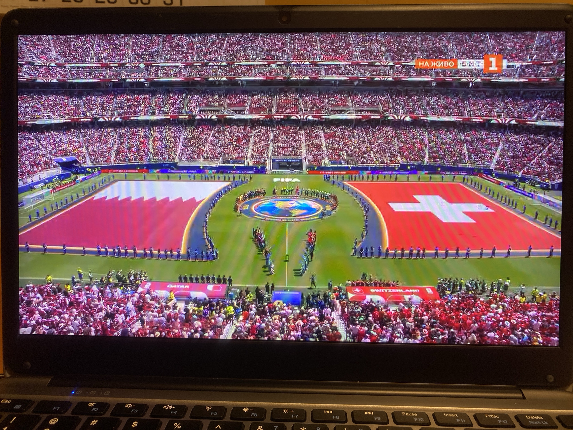 The image displays a large-screen television screen showing a wide shot of a soccer stadium filled with spectators. The field is prominently visible with two large flags, one red and white striped and the other featuring a white cross on a red background. A circular emblem with a depiction of the Earth is in the center of the field, surrounded by a ring of people in uniform. Broadcast graphics are present in the upper right corner, indicating "НА ЖИВО" and a channel number "1". The television is placed on a laptop keyboard, suggesting an indoor viewing environment. The location in the city of Smolyan, Bulgaria is not ascertainable from the visual information presented in the image.