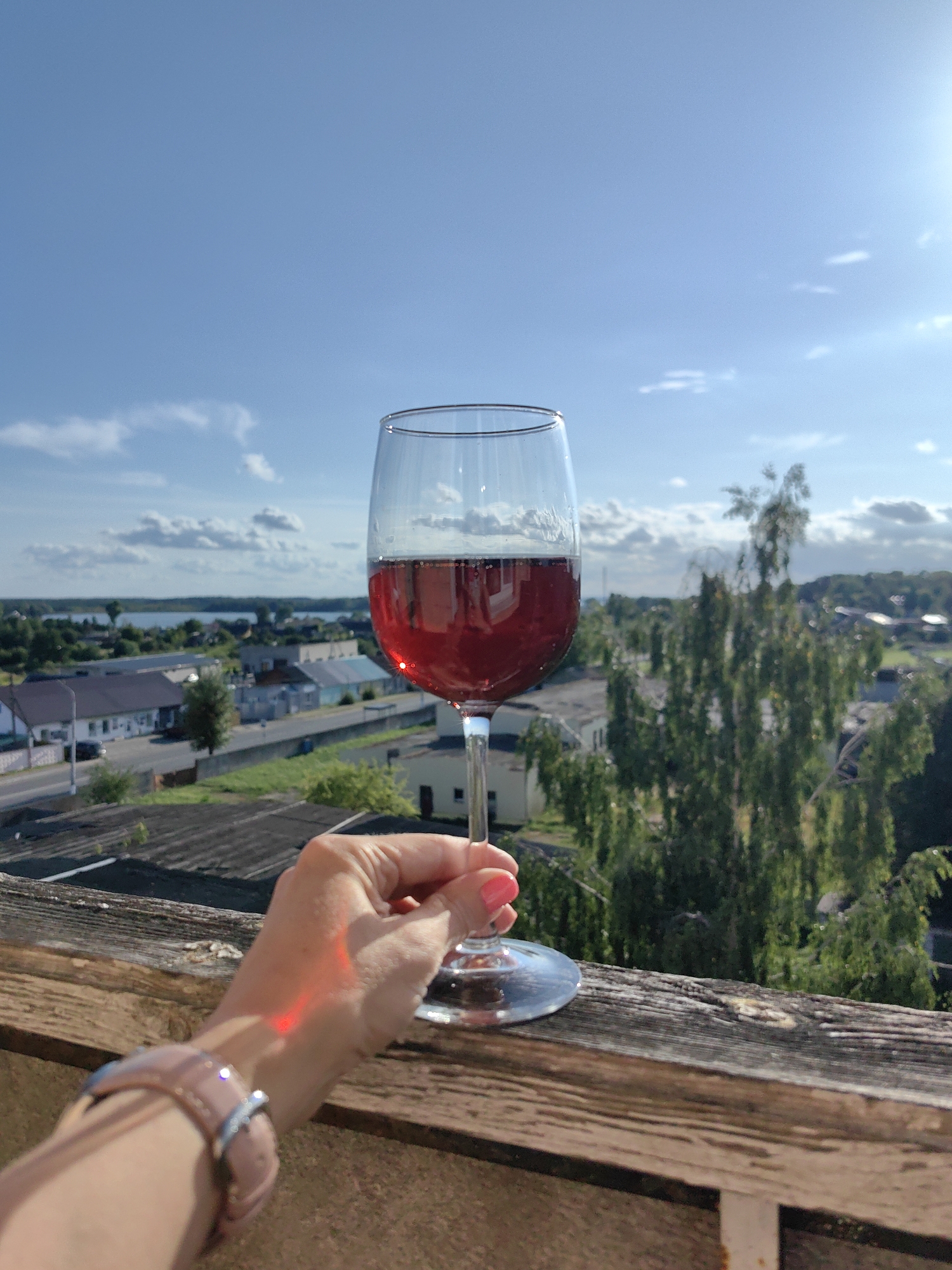 A first-person perspective shot shows a hand holding a wine glass filled with red wine. The hand has bright pink nail polish. A watch with a light-colored band is worn on the wrist. A red light reflection is visible on the back of the hand. The glass is positioned in front of a blurred cityscape and sky. The background shows buildings, trees, and a body of water, likely Lake Myastro, in Myadzyel, Belarus. The sky is blue with scattered white clouds, and the sun is visible on the right side, creating a bright glare. The foreground features a weathered wooden railing.