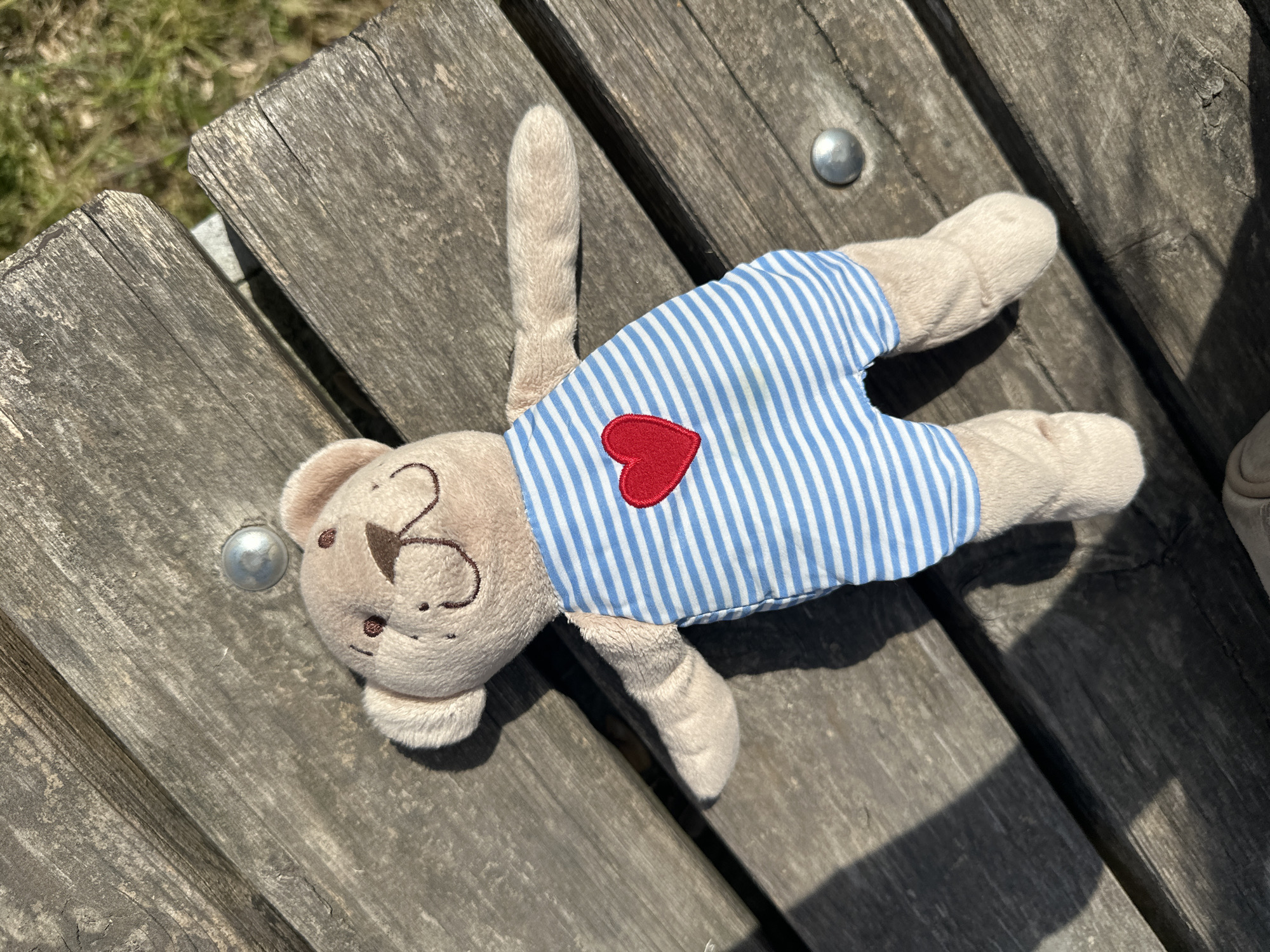A teddy bear wearing a blue and white striped shirt with a red heart is lying on a weathered wooden bench. The bench appears to be outdoors, likely in a park or public space. The lighting suggests it is daytime. Based on the user's prompt, the location is Başakşehir, Turkey.