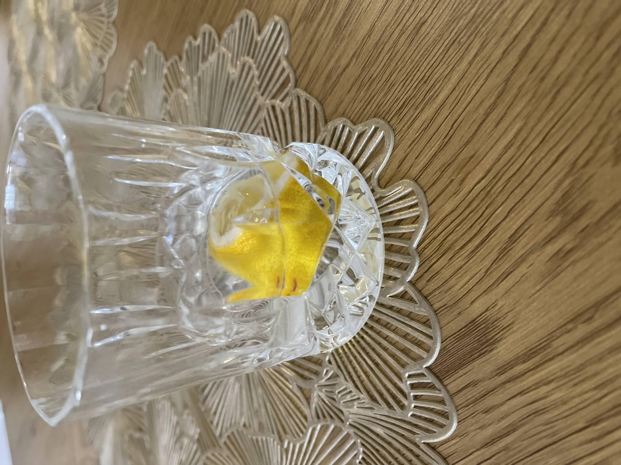 A close-up, eye-level shot captures a crystal glass containing a slice of lemon and some water. The glass is placed on a decorative, metallic placemat with a fan-like pattern, which in turn rests on a wooden table. The background shows a blurred expanse of the placemat and table. The lighting is natural, casting subtle shadows. The scene appears to be indoors, likely in a home or dining setting. There are no people or animals visible in the image. The overall impression is one of simplicity and a focus on the refreshing beverage. The image does not provide any specific visual cues to indicate its location in Podgorica, Montenegro.