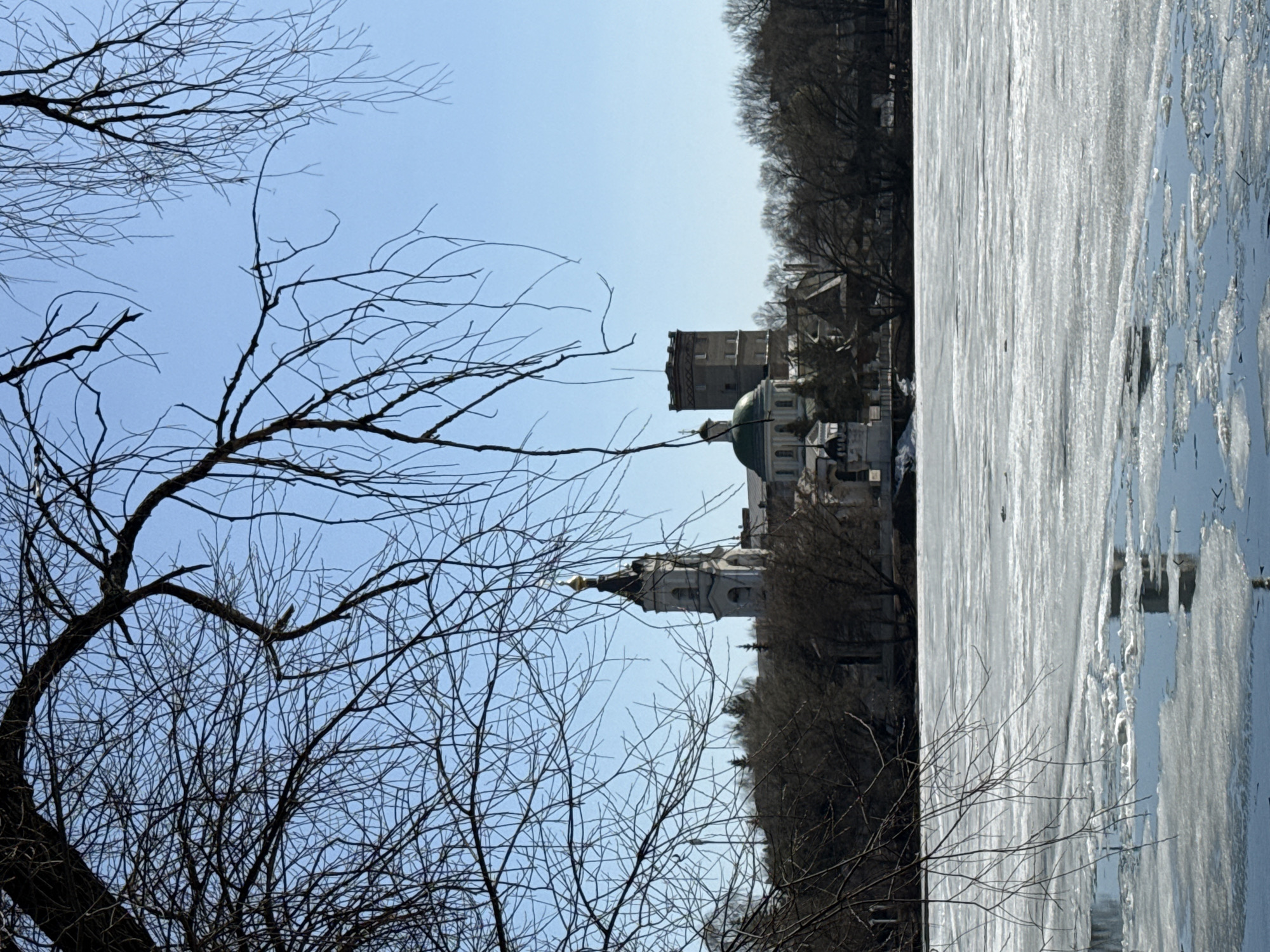 The image shows a winter scene in Moscow, Russia. The foreground is dominated by bare tree branches against a light blue sky. In the midground, the Church of the Holy Martyr Tatiana at Moscow State University and the tower of the Church of the Nativity of John the Baptist can be seen. In the background is a frozen body of water, likely a pond or part of the Moscow River, with visible ice formations. No people are visible in the image. The environment suggests a cold, early spring day in an urban park or near the university campus.