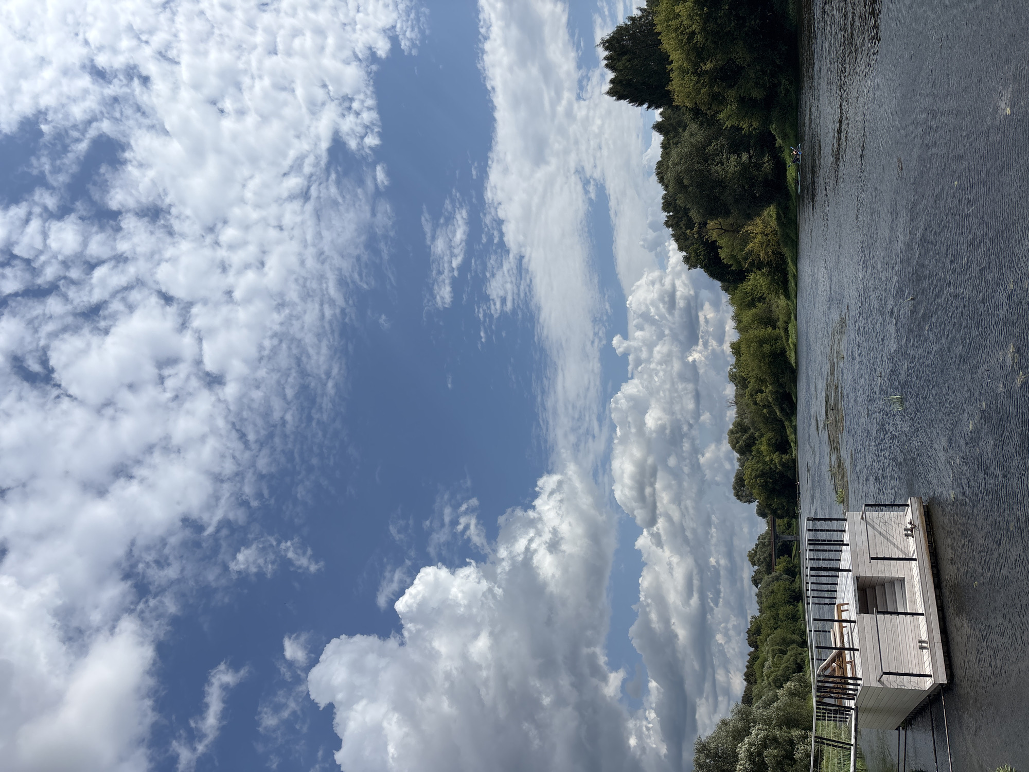 The image depicts a serene river scene on a partly cloudy day, likely in Zvenigorod, Russia, given the context. In the foreground, a modern, white wooden floating structure is moored to the left side of the frame. This structure has a deck with railing and two lounge chairs. Steps lead down from the deck to the water. The river itself is calm with ripples on its surface, and patches of aquatic vegetation are visible.
The riverbanks are lined with lush green trees, forming a dense forest. In the distance, a bridge can be seen spanning the river. The sky is a vibrant blue, dotted with numerous white, fluffy clouds, some of which are quite large and imposing, suggesting dynamic weather patterns. To the right, a small boat with two occupants is visible on the water. The overall atmosphere is peaceful and natural.