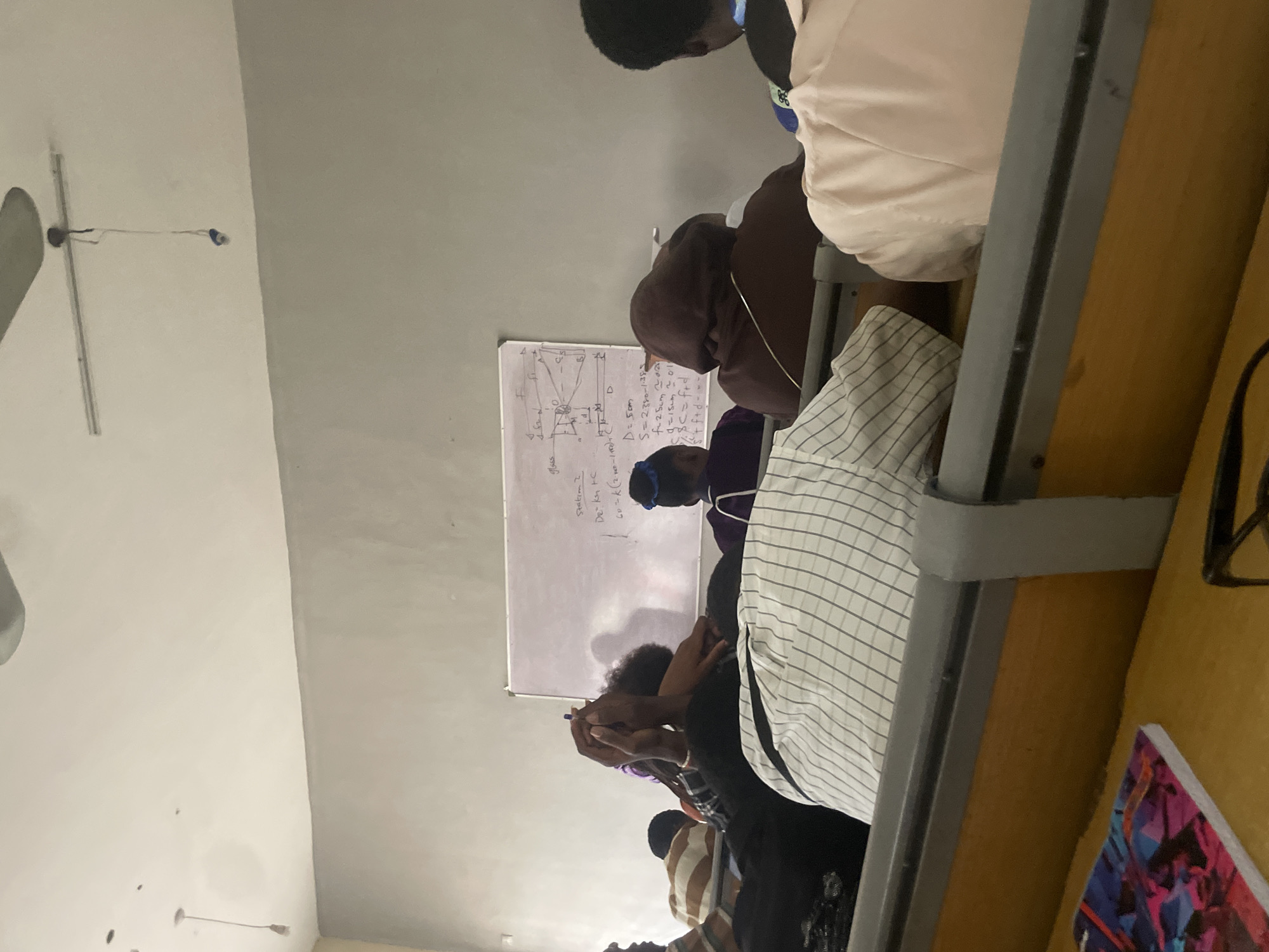 This is a medium shot photograph taken from behind students seated in a classroom in Ihiagwa, Nigeria. The room is dimly lit with a bare light bulb hanging from the ceiling. A whiteboard is visible at the front of the room, covered in handwritten equations and a diagram that appears to be related to optics. Several students are facing away from the camera, their backs to the viewer, indicating they are likely attending a lecture or lesson. Some students are wearing hoodies, and one student in the foreground has their arms crossed and head down. The classroom furniture consists of wooden desks with metal framing. A notebook with a colorful abstract design is on a desk in the foreground. The overall atmosphere suggests a typical academic setting.