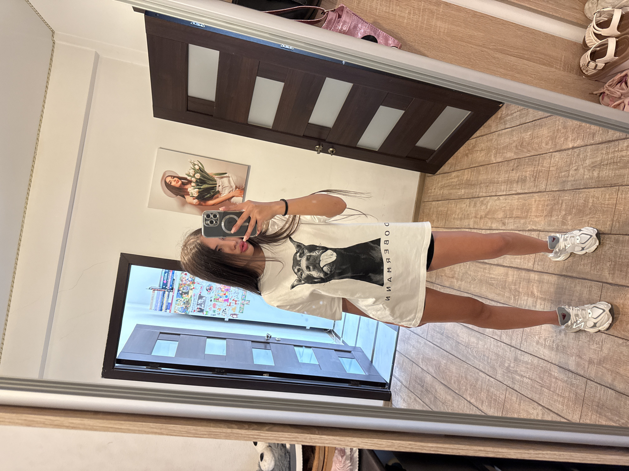 A young woman stands in a hallway, taking a mirror selfie. She is wearing an oversized white t-shirt with a graphic of a Doberman Pinscher on it, black shorts, and white sneakers. She has long, dark hair and is wearing subtle makeup. The hallway features wooden floors and doors, and a framed picture of a woman holding flowers hangs on the wall. The scene appears to be within a residential building.