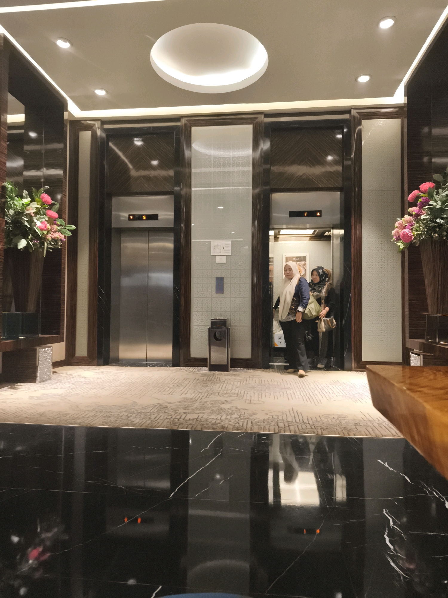 The image depicts an interior scene, likely in a hotel lobby or a similar establishment. Two elevators are visible. The elevator on the left has its doors fully closed, with the number "5" displayed on its indicator. The elevator on the right has its doors open, and two women are seen inside. One woman, wearing a blue hijab and jacket, stands facing forward, while the other, in a patterned hijab and dark clothing, stands behind her, looking towards the front. They both appear to be waiting or have just entered the elevator.
The environment is well-lit with modern lighting fixtures on the ceiling, including a circular cove light. The walls feature dark wood paneling and patterned wallpaper. A patterned carpet covers the floor in the foreground, which transitions to a polished black marble floor with white veining. A vase filled with pink and white flowers is placed to the left of the left elevator, and another floral arrangement is visible to the right of the right elevator. A trash receptacle is situated near the right elevator. The overall ambiance suggests a sophisticated and contemporary setting. The presence of elevators and the luxurious decor are indicative of a hospitality or commercial building. The specific location in Palembang, Indonesia, is not explicitly indicated by any visual cues in the image.
