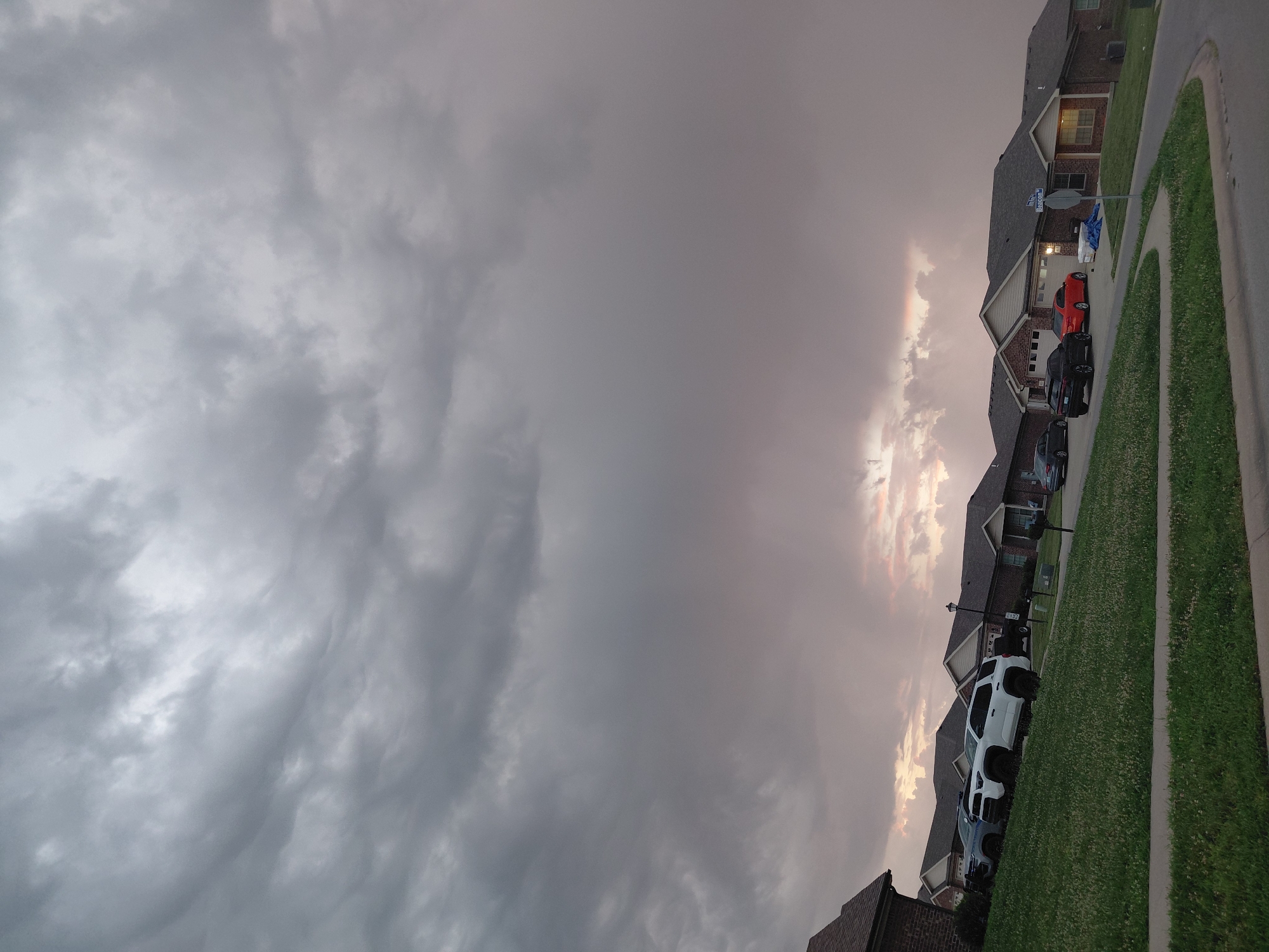 This is a photograph taken at street level, looking across a suburban street toward a row of houses. The sky is heavily overcast with dark, textured clouds, suggesting an impending storm or recent rain. The lighting is dim, casting a somber mood over the scene. In the foreground, a well-manicured lawn slopes upward towards the houses. Several vehicles are parked in the driveways and on the street. The houses themselves appear to be single-family residences, constructed of brick with dark-colored roofs. A street sign visible on the right side of the image reads "Beacon." The overall setting suggests a residential neighborhood in Richmond, United States of America, likely during the late afternoon or early evening given the diffused light.