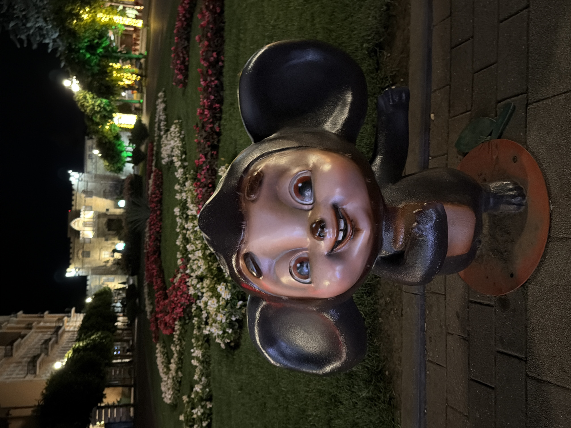 A statue of a cartoon mouse character with large ears, dark brown and pink coloring, and a wide smile stands in the foreground on a paved walkway. Behind the statue, a garden with neatly trimmed grass and beds of red and white flowers is illuminated by artificial lights. In the background, a building with ornate architecture is visible, as are trees decorated with colorful string lights. The scene appears to be a public park or square in Kislovodsk, Russia, at night.